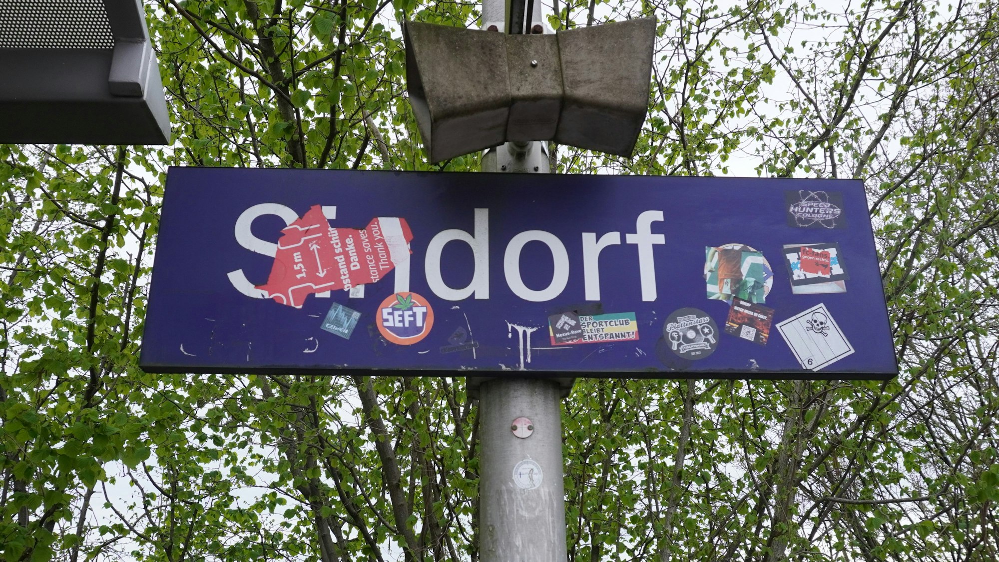 Das Foto zeigt ein S-Bahn-Haltestellenschild mit der Aufschrift „Sindorf“, das teils überklebt wurde.
