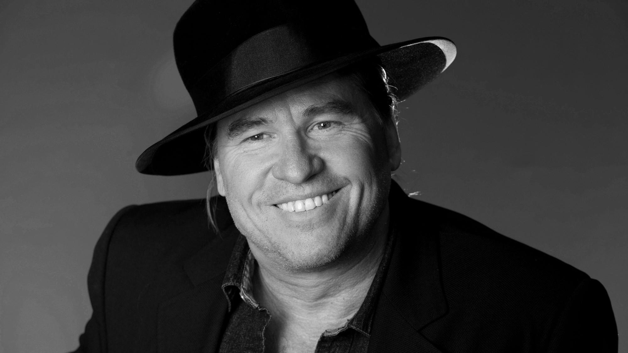 Im Schwarz-Weiß-Porträt lächelt Val Kilmer in die Kamera. Er trägt einen großen schwarzen Hut.