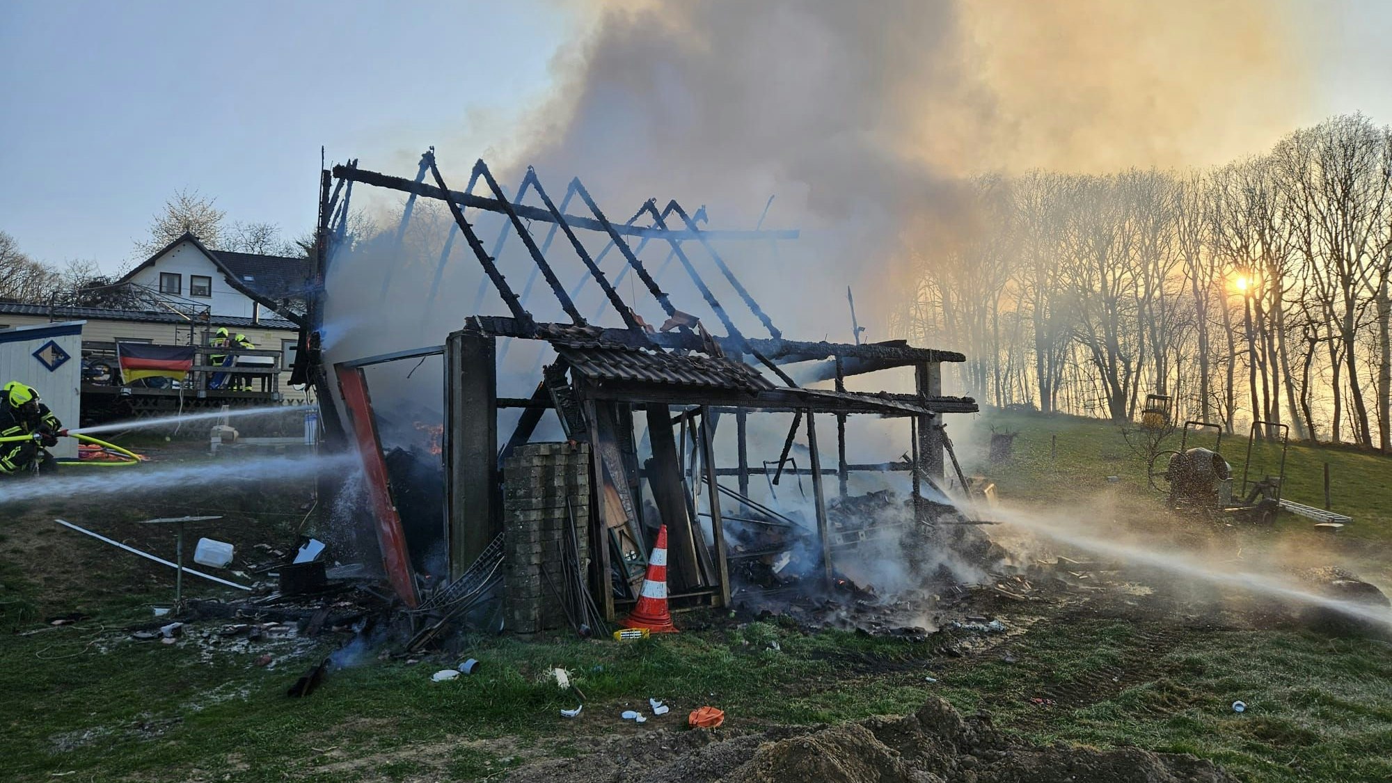 Eine abgebrannte Scheune, aus der Qualm kommt. Links ein Wasserstrahl und Einsatzkräfte der Feuerwehr.