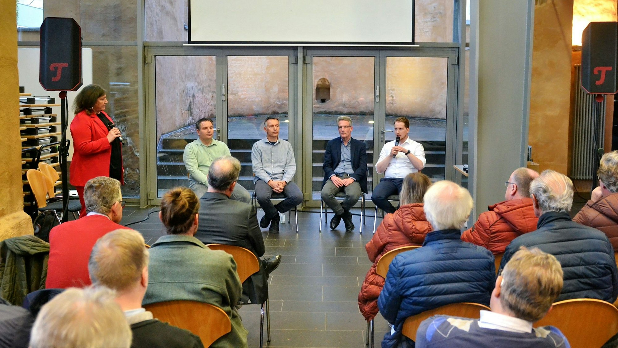Vor einem Publikum, das auf Stühlen sitzt, stehen eine Frau und vier Männer sitzen auf Stühlen.