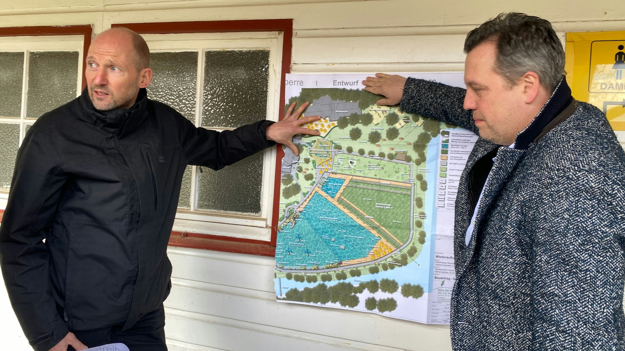 Zwei Männer halten einen Plan des sanierten Freibades an eine Holzwand.