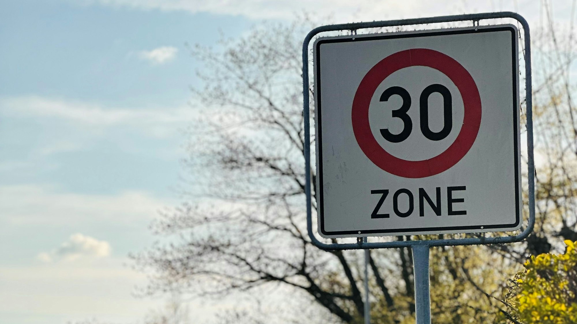 Zu sehen ist ein Tempo 30 Schild vor blauem Himmel und Bäumen.