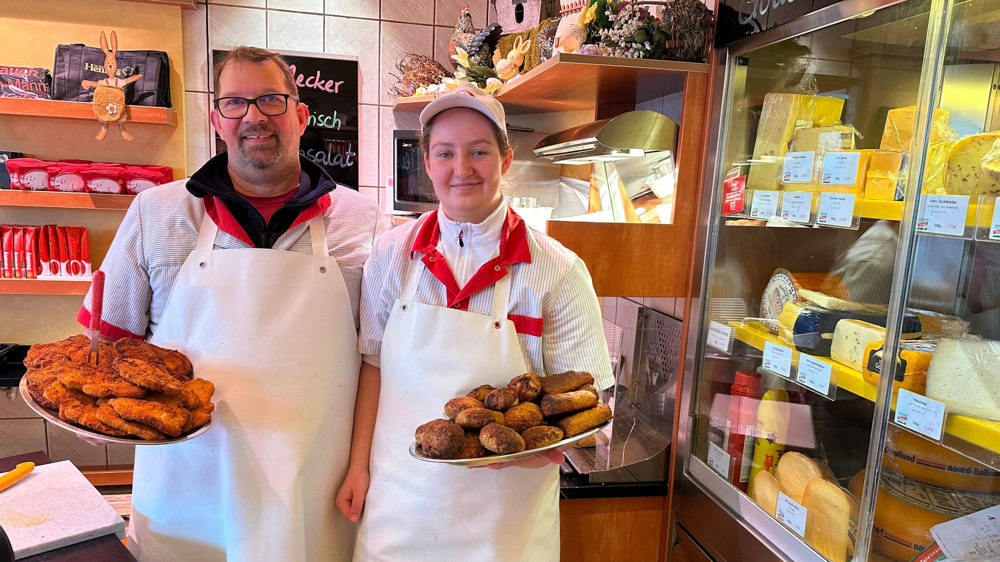 Ein Mann und eine Frau stehen nebeneinander in einer Metzgerei. Beide halten Teller in der Hand, auf dem einen sind Schnitzel, auf dem anderen Frikadellen.