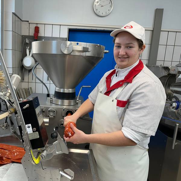 Eine junge Frau in Metzger-Schürze und mit Käppi auf dem Kopf steht an der Wurstmaschine.