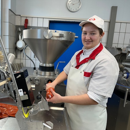 Eine junge Frau in Metzger-Schürze und mit Käppi auf dem Kopf steht an der Wurstmaschine.