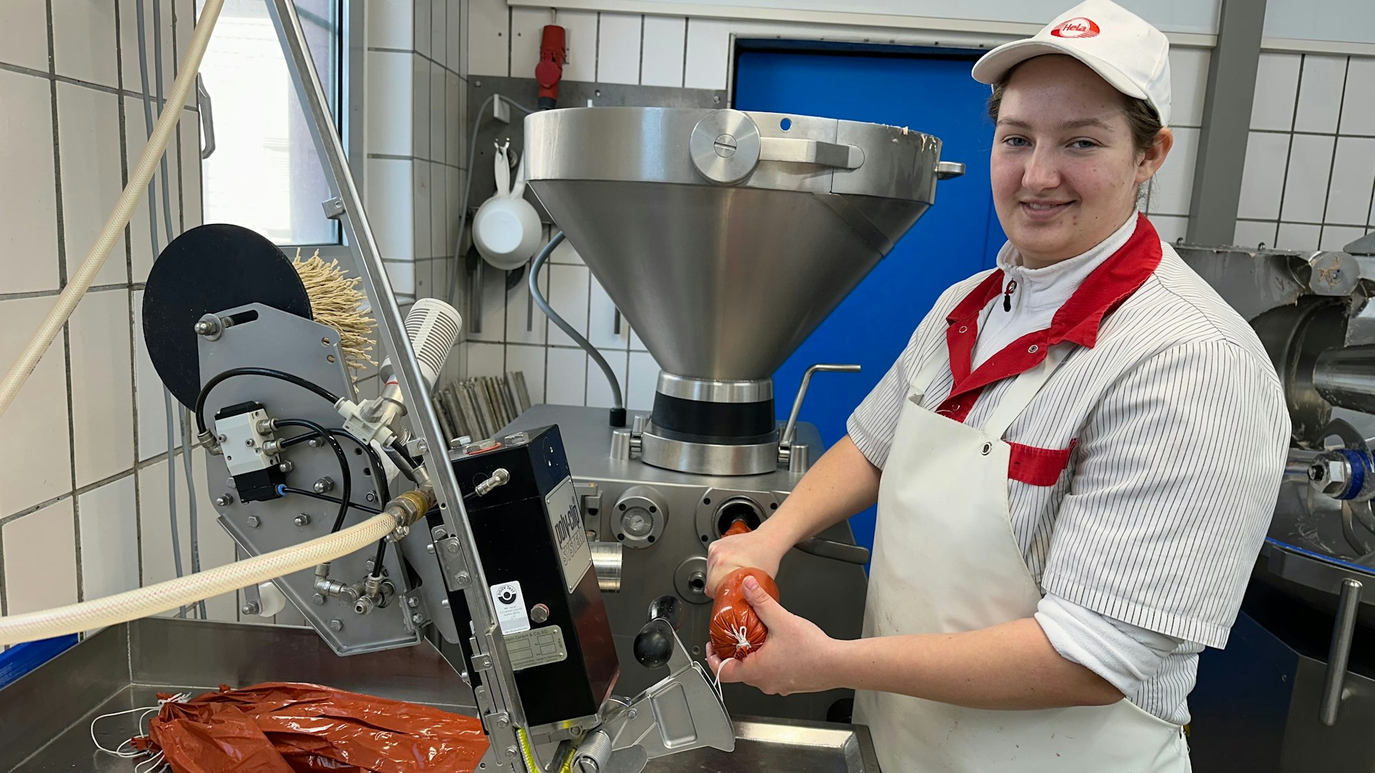 Eine junge Frau in Metzger-Schürze und mit Käppi auf dem Kopf steht an der Wurstmaschine.