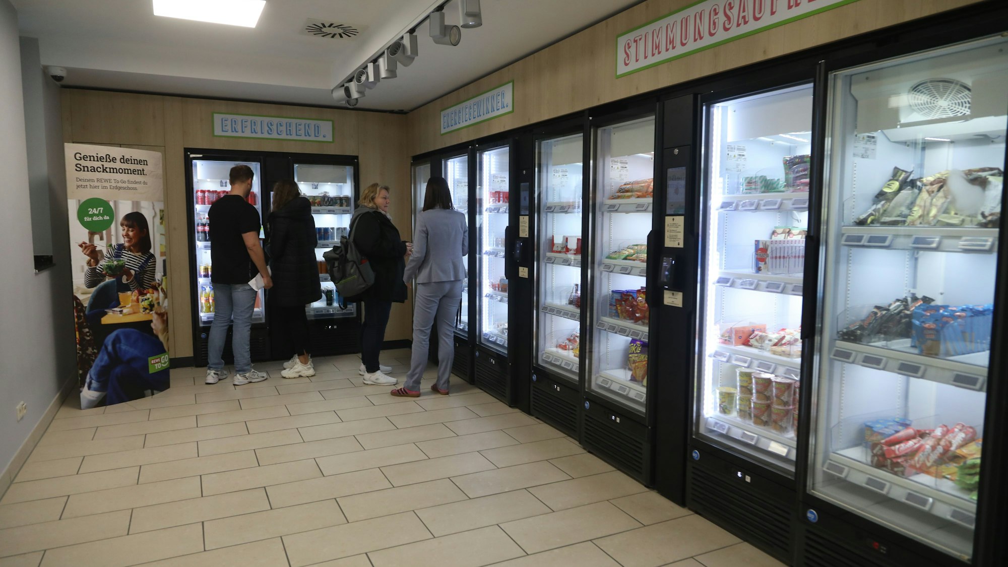 In einem Verkaufsraum stehen Kühlschränke, gefüllt mit Snacks und Schokolade. Menschen stehen davor und suchen sich etwas aus.