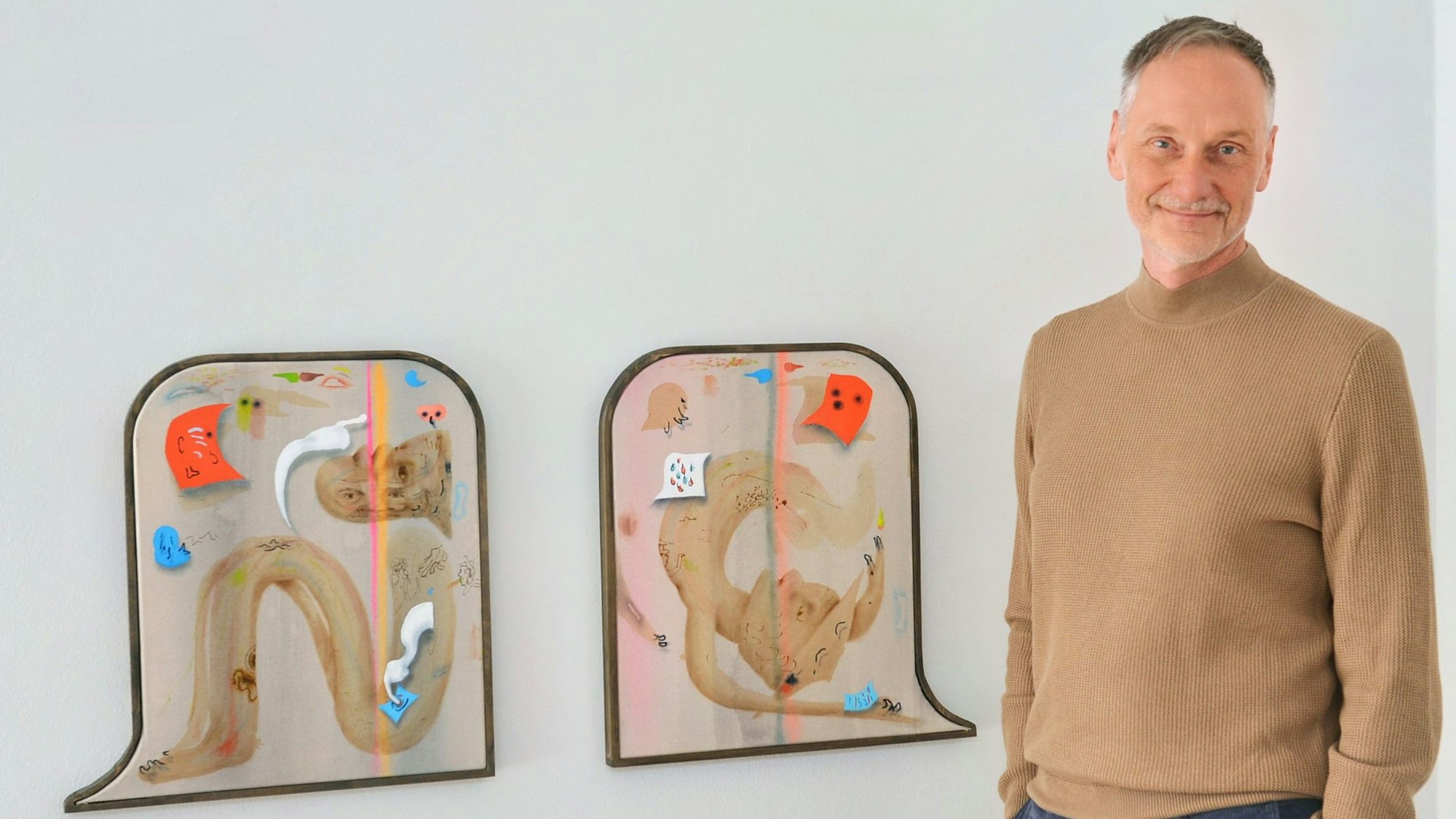 Ein Mann in einem braunen Rollkragenpullover steht neben zwei Bildern. Die Werke sind vollgepackt mit unterschiedlichen Details.