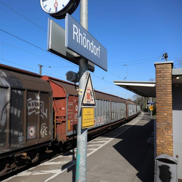 Ein Schild mit dem Schriftzug Rhöndorf auf einem Gleis des Bahnhofs, durch den ein Güterzug fährt.