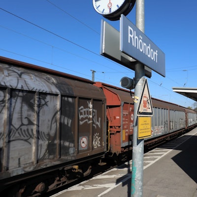 Ein Schild mit dem Schriftzug Rhöndorf auf einem Gleis des Bahnhofs, durch den ein Güterzug fährt.