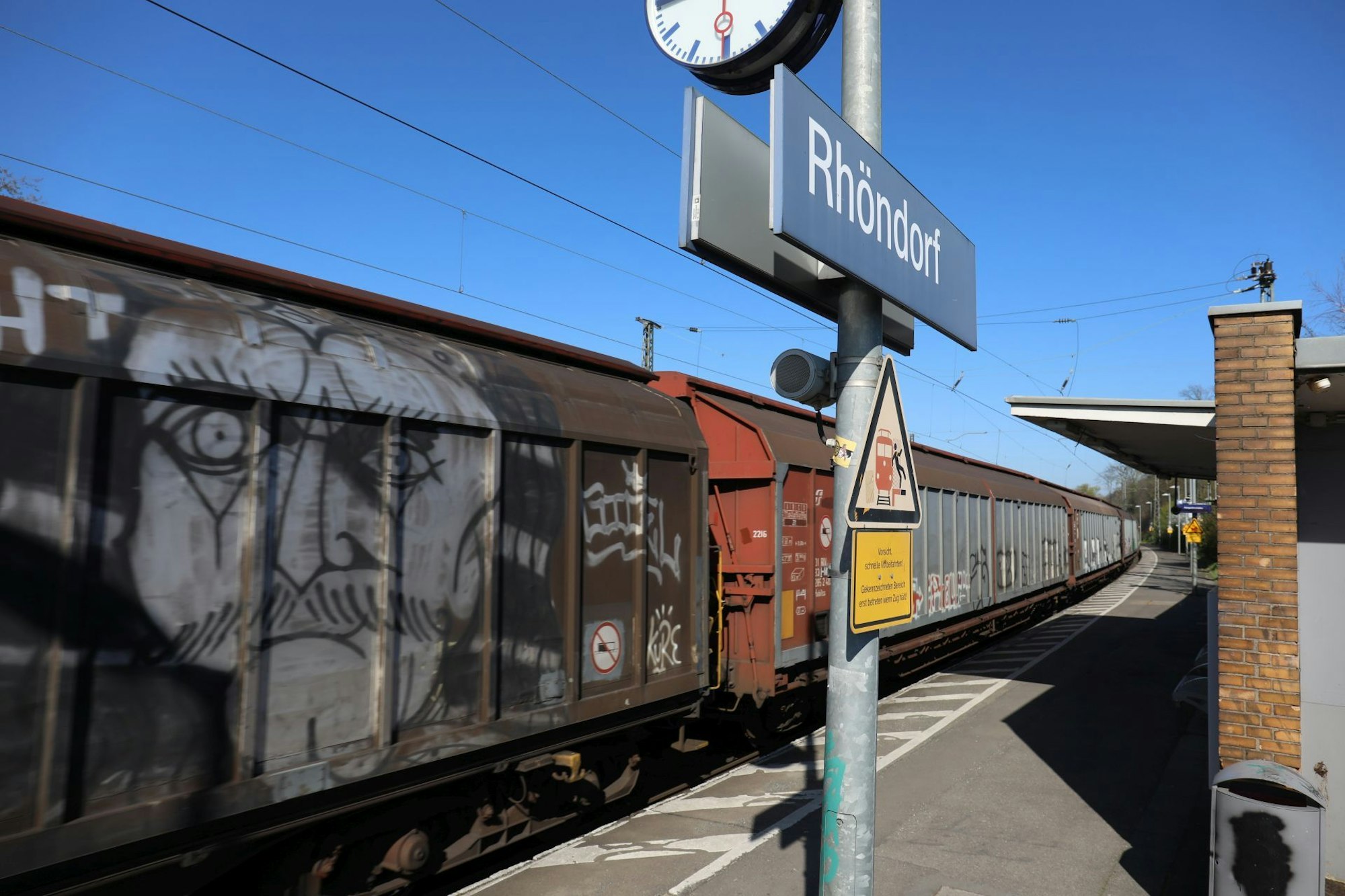 Ein Schild mit dem Schriftzug Rhöndorf auf einem Gleis des Bahnhofs, durch den ein Güterzug fährt.