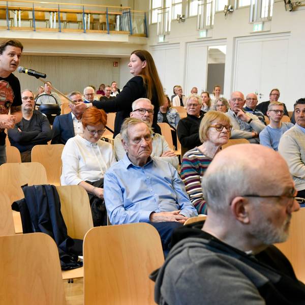 Mehrere Menschen sitzen in Stuhlreihen, ein Mann spricht in ein Mikrofon, das ihm eine Frau hinhält.