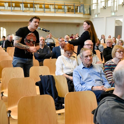 Mehrere Menschen sitzen in Stuhlreihen, ein Mann spricht in ein Mikrofon, das ihm eine Frau hinhält.