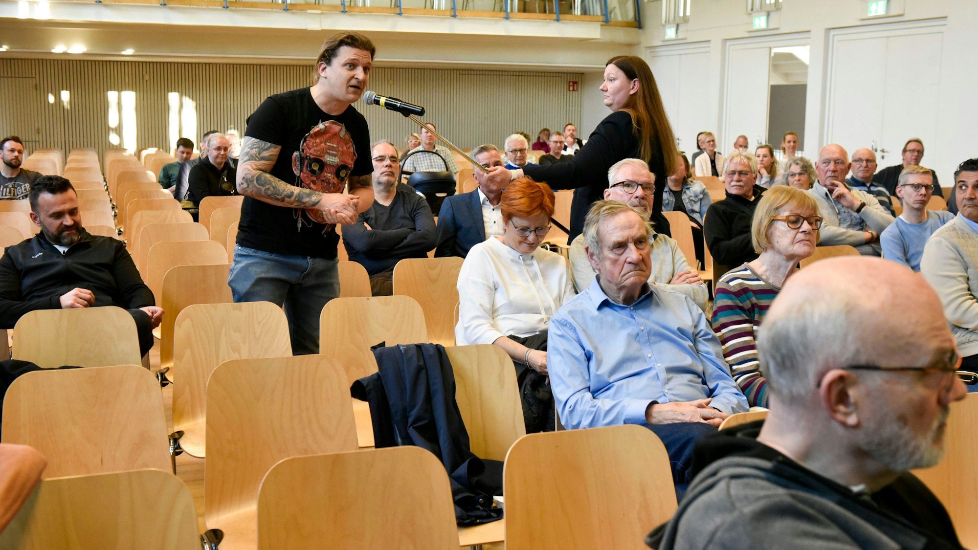 Mehrere Menschen sitzen in Stuhlreihen, ein Mann spricht in ein Mikrofon, das ihm eine Frau hinhält.