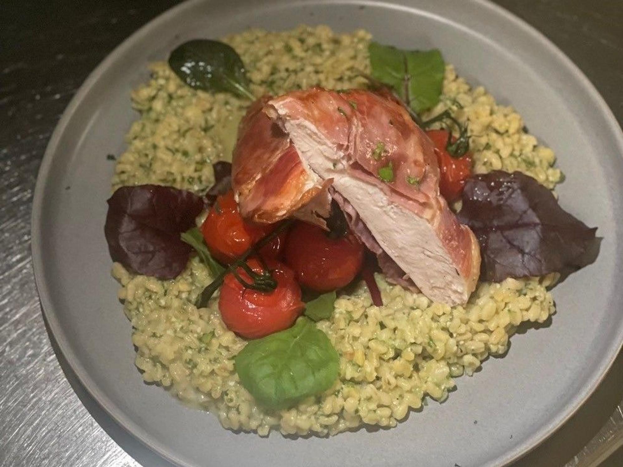 Zu sehen ist ein Teller Graupenrisotto mit Bärlauch, Cherrytomaten, Wildkräutersalat, Hähnchenbrust und Serranoschinken