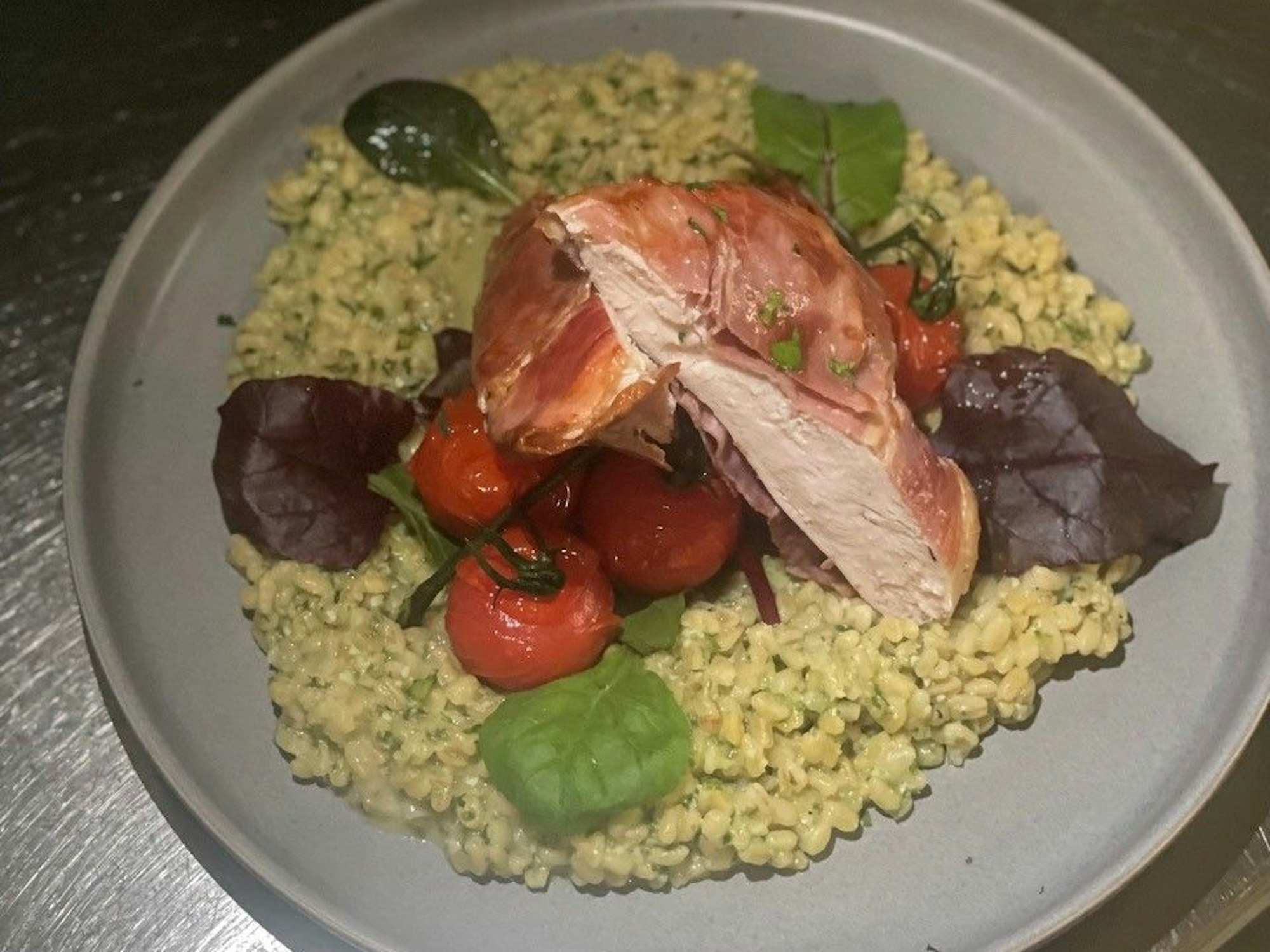 Zu sehen ist ein Teller Graupenrisotto mit Bärlauch, Cherrytomaten, Wildkräutersalat, Hähnchenbrust und Serranoschinken