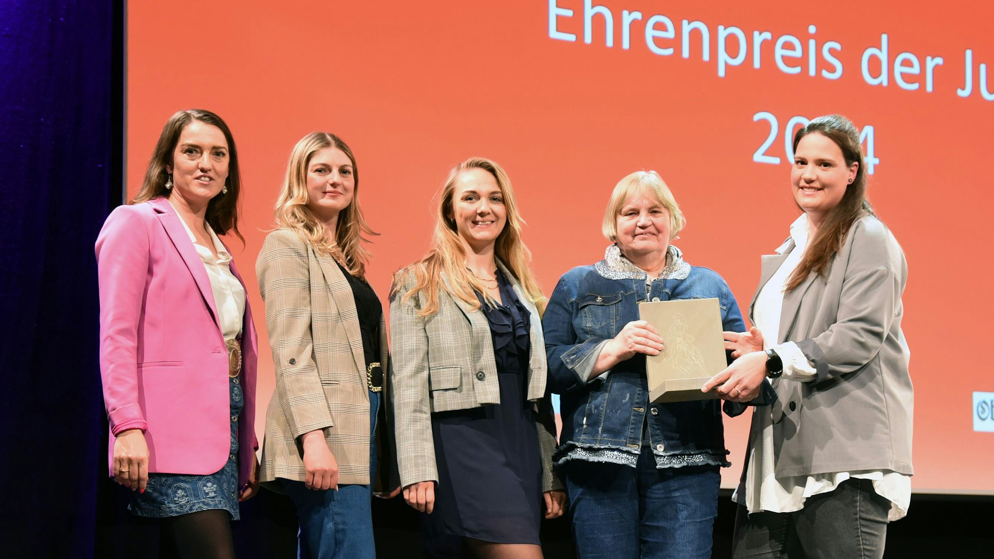 Fünf Frauen stehen auf einer Bühne nebeneinander. Eine Frau überreicht einer anderen eine Skulptur aus Grauwacke.