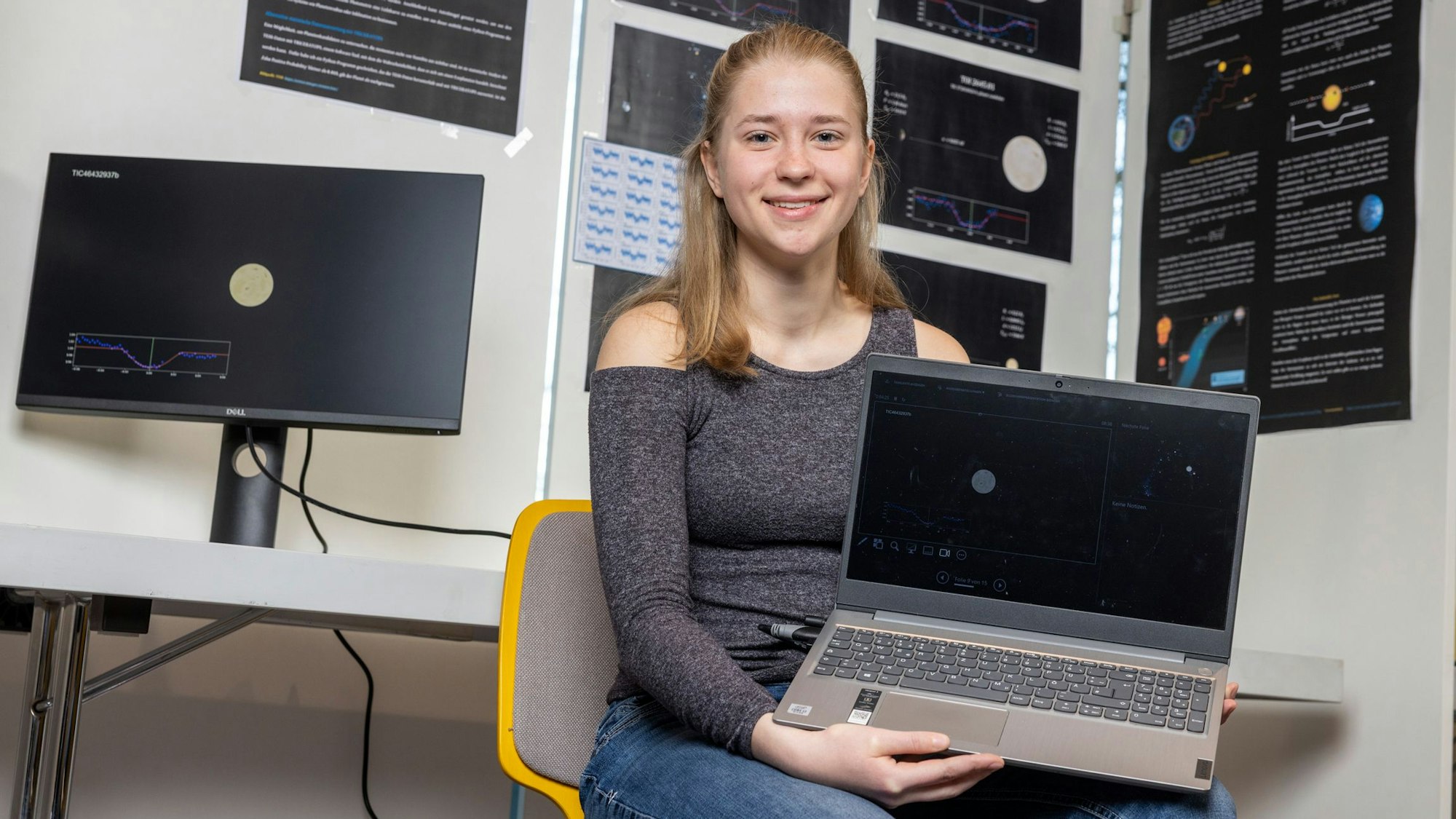 Emily Hein sitzt vor einer Wand mit vielen Bildern zum Thema Weltall und hält einen Laptop in den Händen.