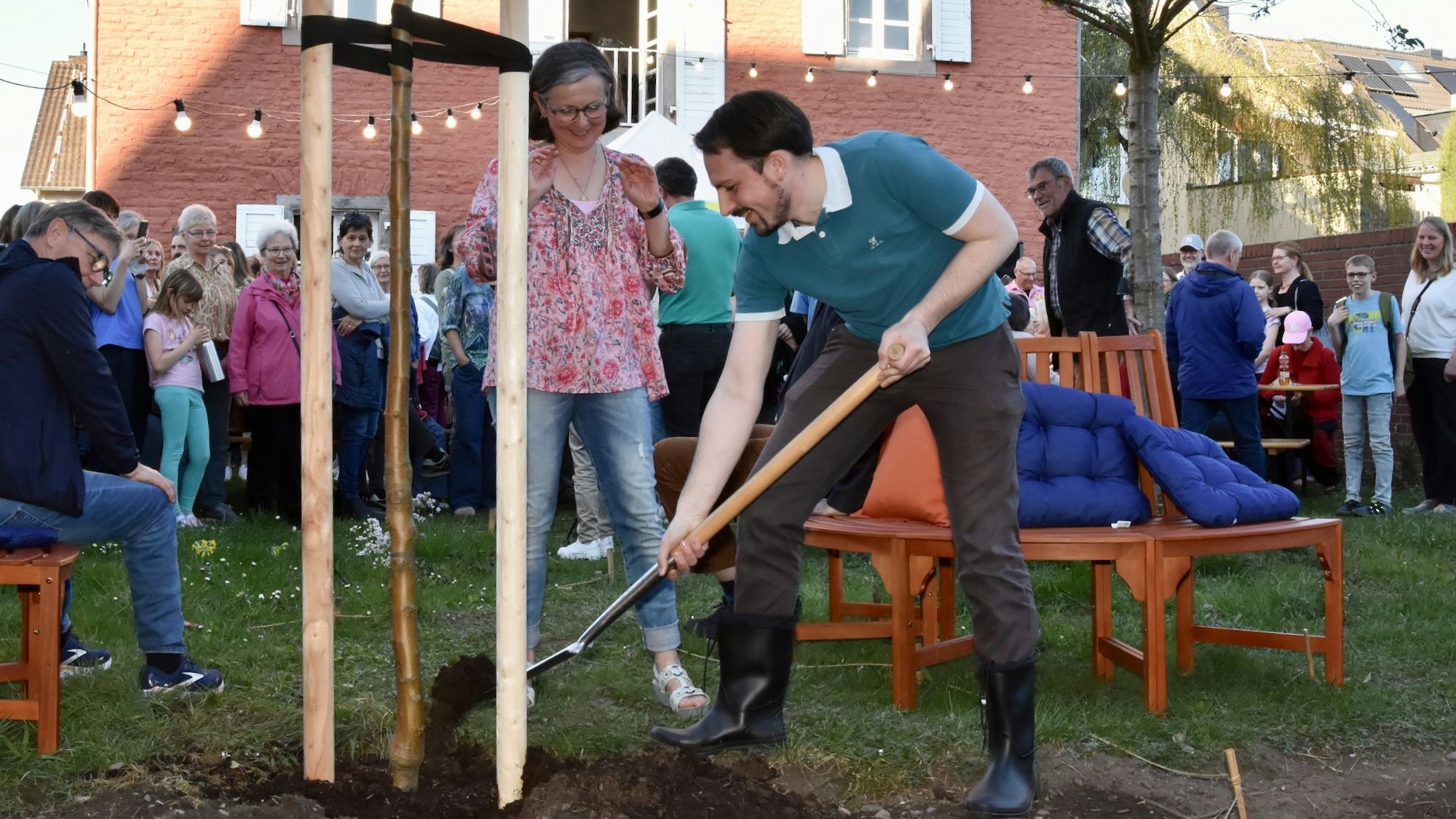 Ein Mann und eine Frau pflanzen einen Baum in einem Garten. Ringsum stehen Menschen und schauen zu.