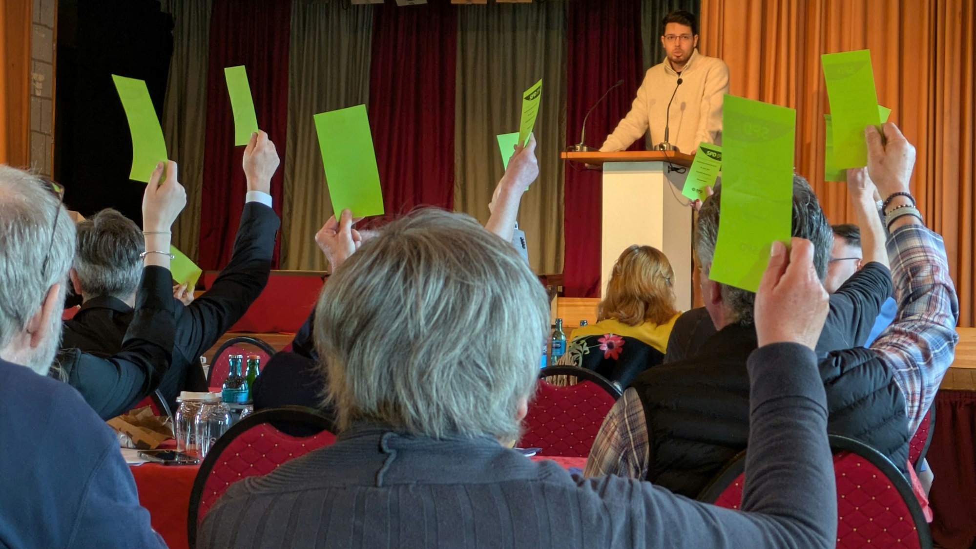 Ein Mann steht an einem Rednerpult, das Publikum hält grüne Karten hoch.