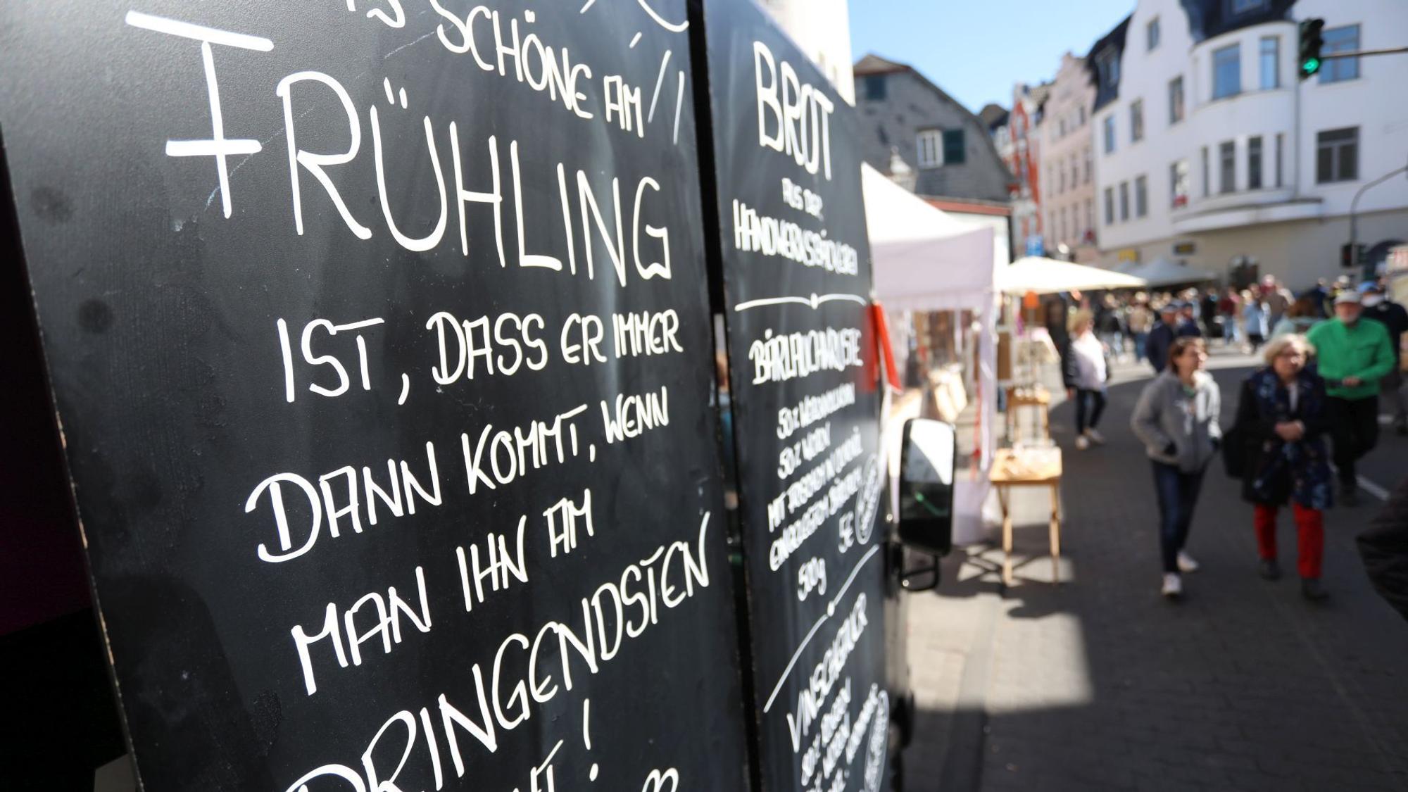 Ein Schild mit einem Text zum Frühling, im Hintergrund Marktstände und Festbesucher.