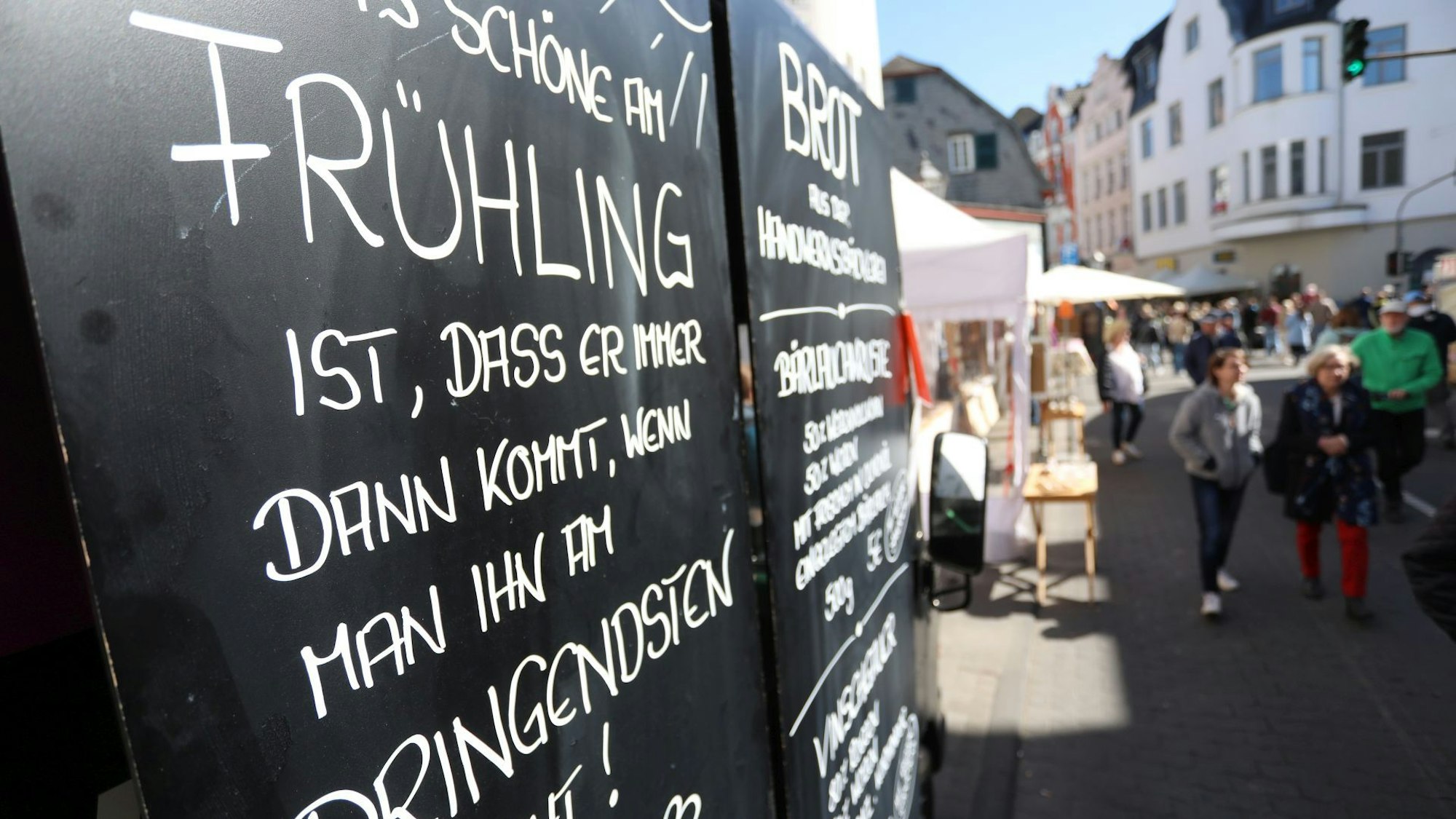 Ein Schild mit einem Text zum Frühling, im Hintergrund Marktstände und Festbesucher.
