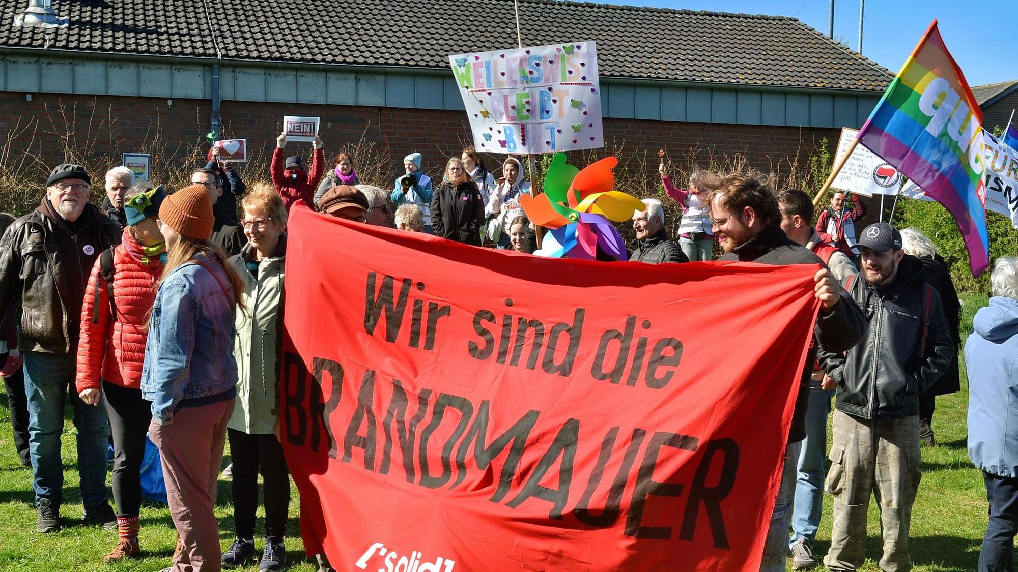 Das Bild zeigt Demonstrierende. Zwei von ihnen halten ein Banner mit der Aufschrift „Wir sind die Brandmauer“.