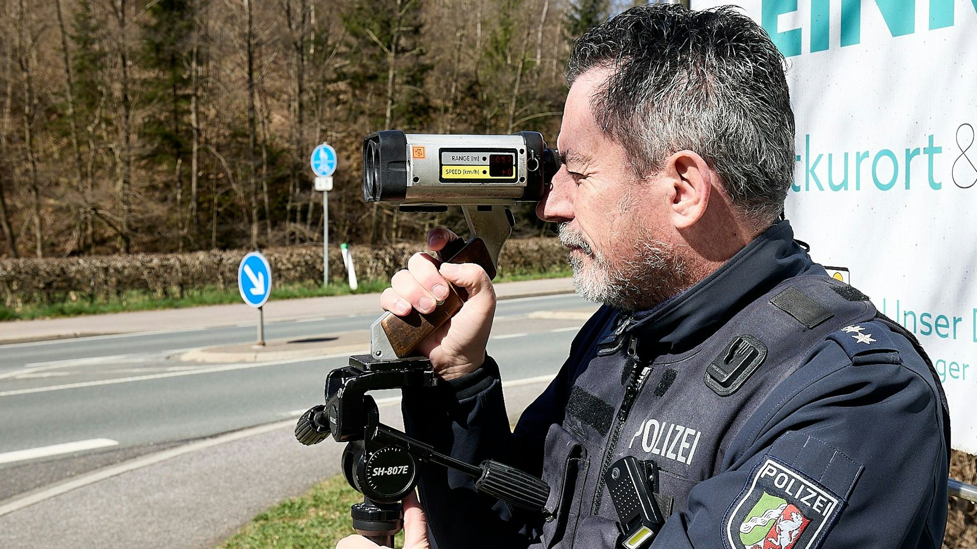Ein Polizist steht an einer Straße in der Eifel und misst mit einem Lasergerät die Geschwindigkeit der vorbeifahrenden Autos und Motorräder.