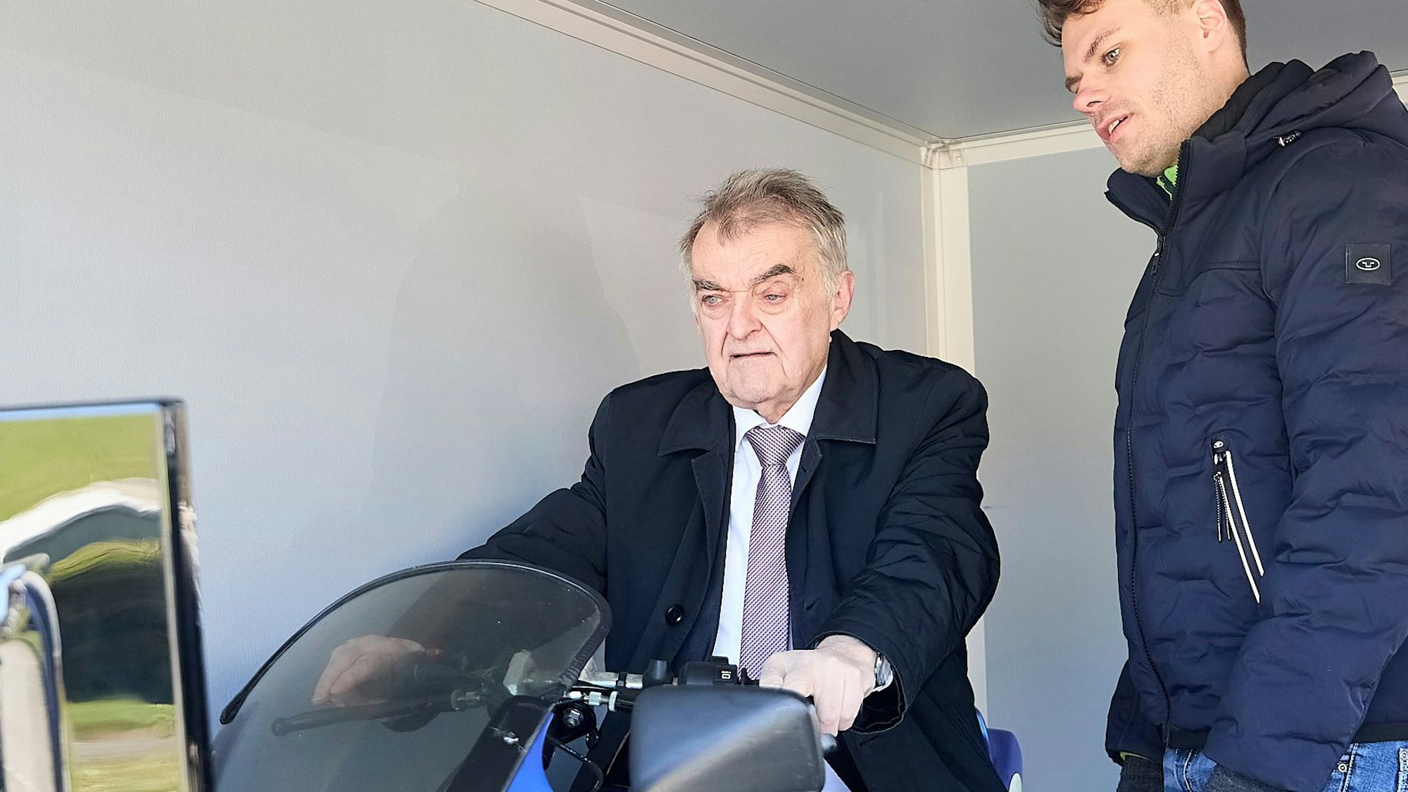 NRW-Innenminister Herbert Reul sitzt auf einem Motorrad-Simulator. Ein Mann schaut ihm dabei zu.