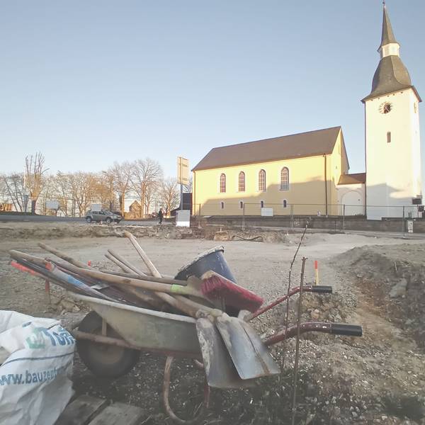 Vorn eine Schubkarre mit Werkzeug, hinten eine Kirche.