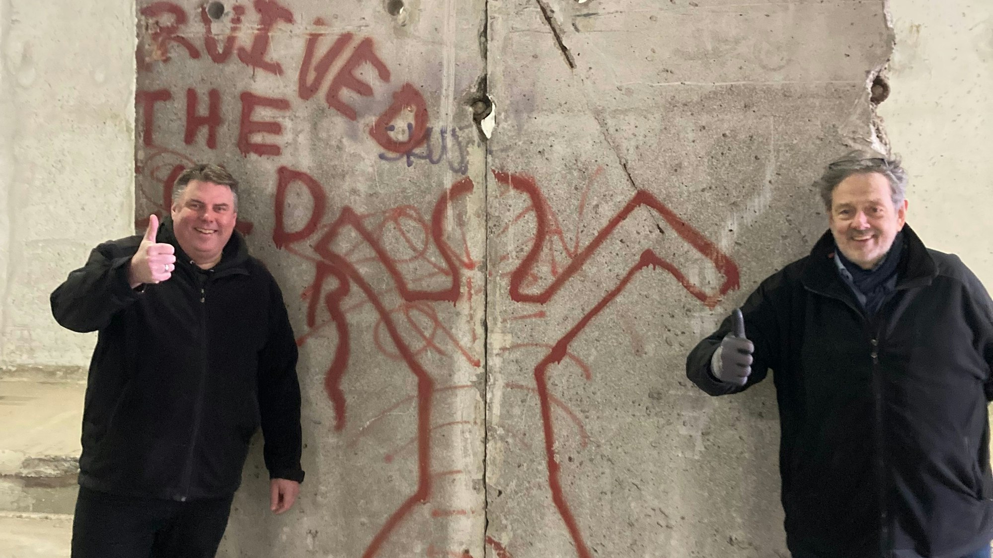 Zwei Männer in schwarzen Jacken stehen an einem Segment der Berliner Mauer. Sie jecken jeweils einen Daumen nach oben.
