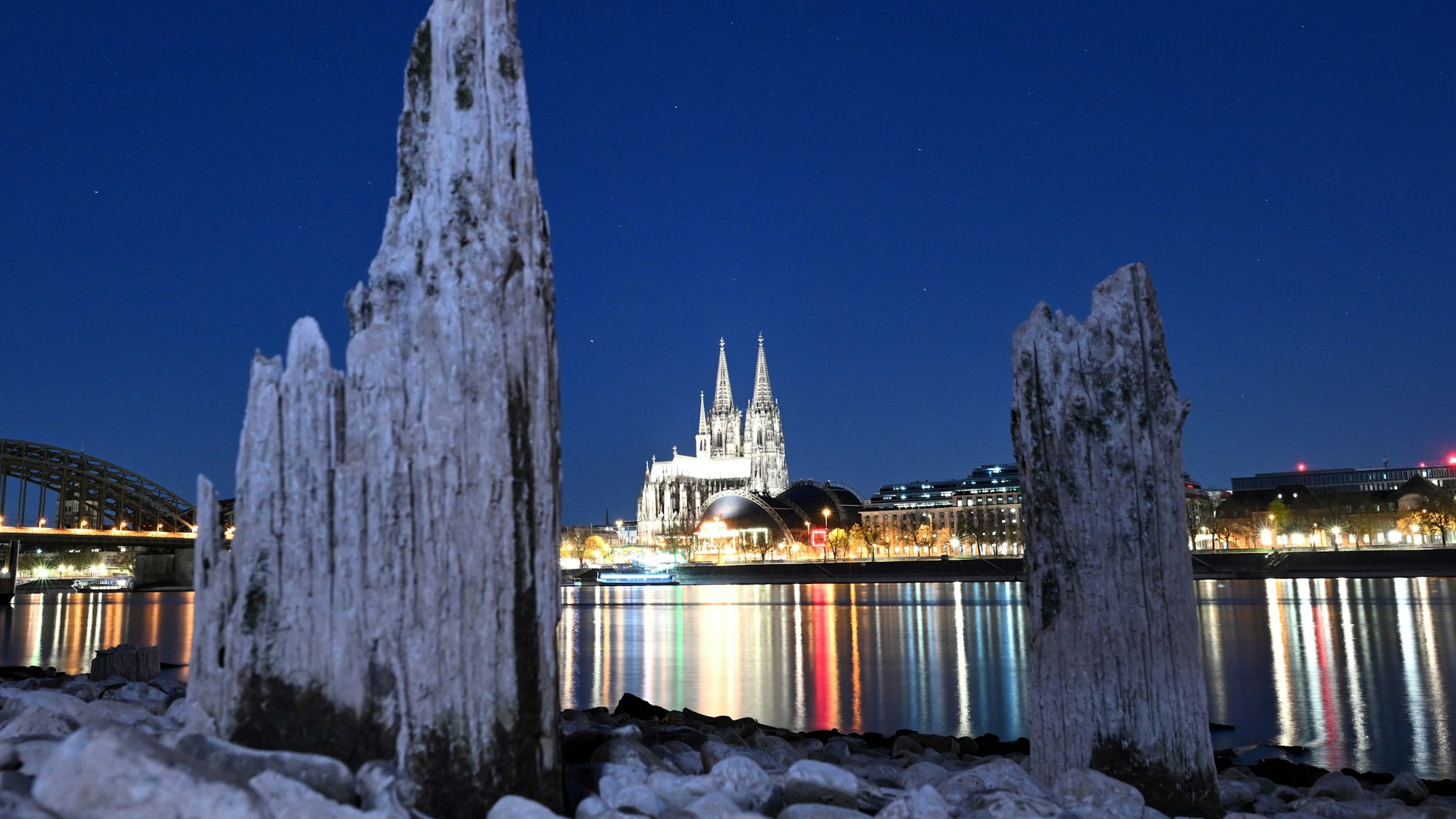 Teile einer Holzkonstruktion, die normalerweise unter Wasser liegen, sind in der Nacht zum Montag (7. April) am Rhein zu sehen. Der Kölner Dom im Hintergrund.