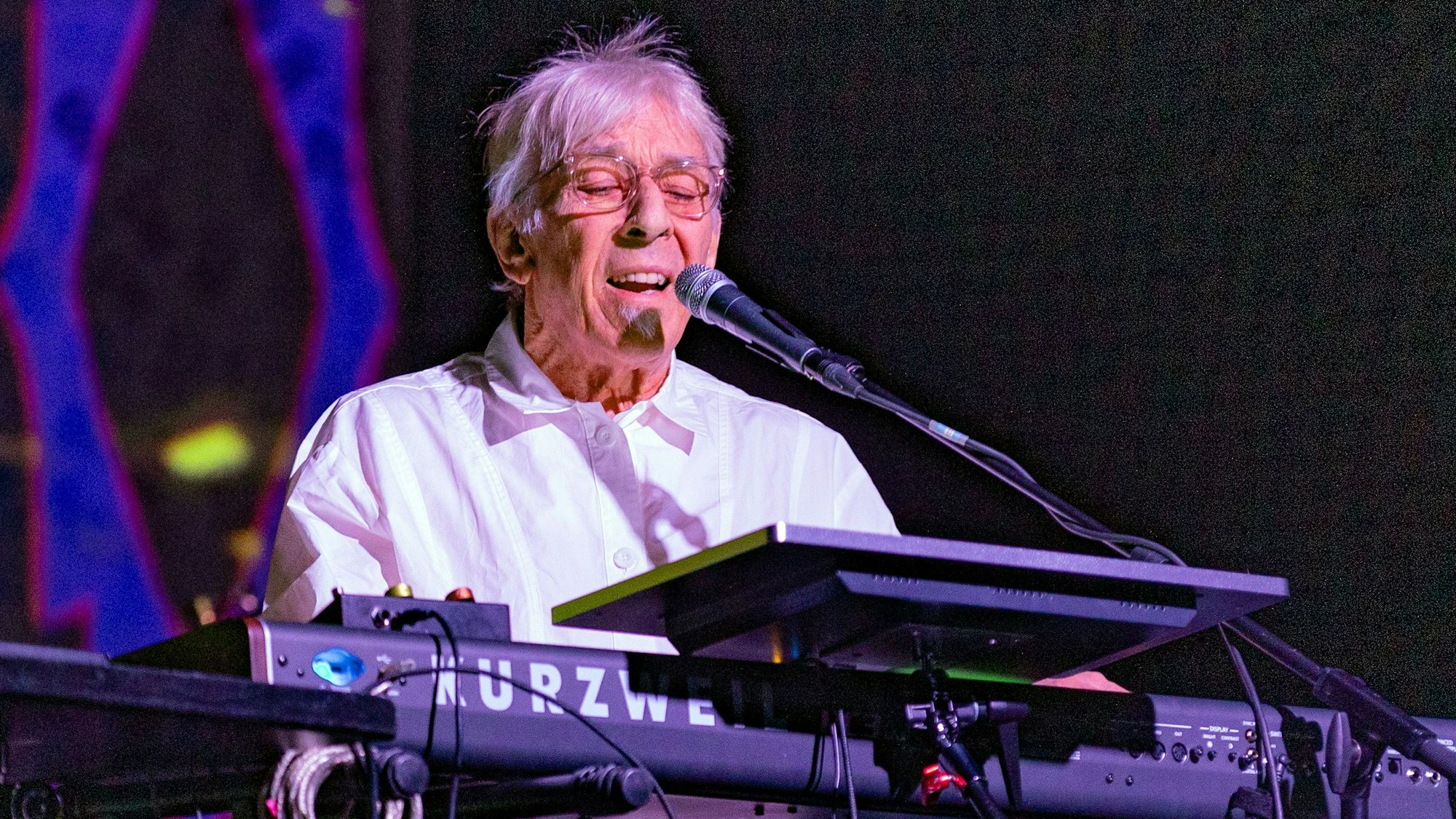 John Cale spielt Keyboard und singt auf der Bühne des Carlswerk Victoria. Er trägt ein weißes Hemd und Brille.