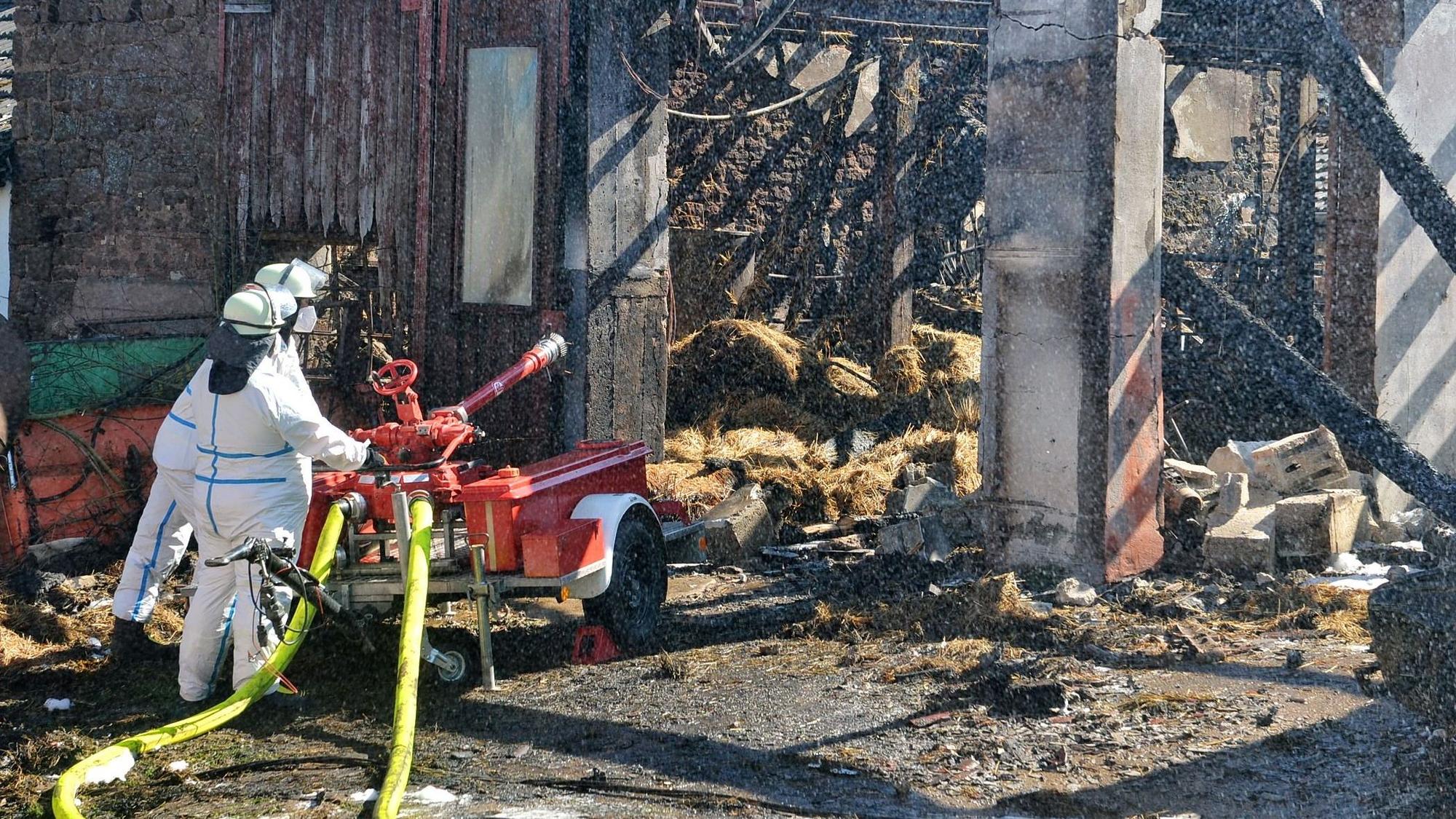 Zwei Feuerwehrleute in weißen Schutzanzügen stehen an einem Wasserwerfer, der auf die Trümmer der niedergebrannten Halle gerichtet ist.