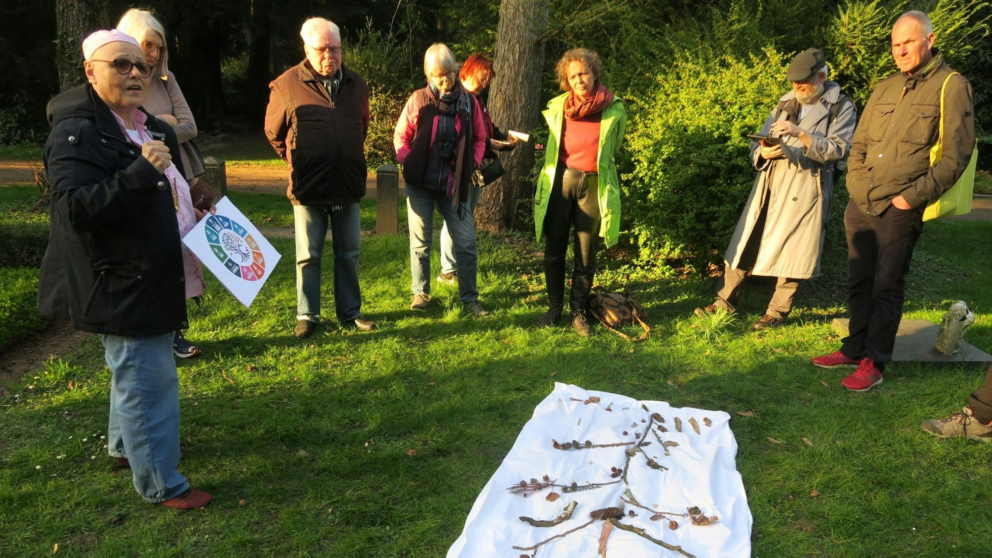 Menschen stehen im Halbkreis auf einem Stück Rasen, auf einem Tuch haben sie Zweige und Blätter zu einem Baum angeordnet.