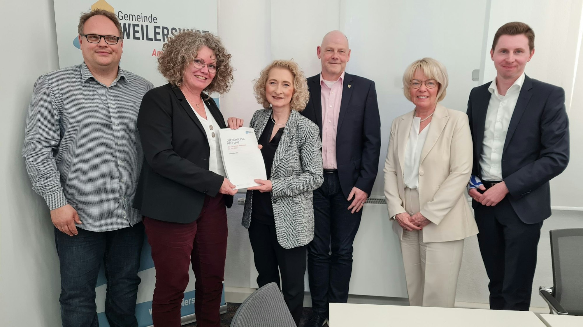 Der Bürgermeisterin Anna-Katharina Horst wird ein Bericht überreicht. Sie und fünf weitere Personen stehen in einem Raum der Gemeinde Weilerswist und schauen in die Kamera.