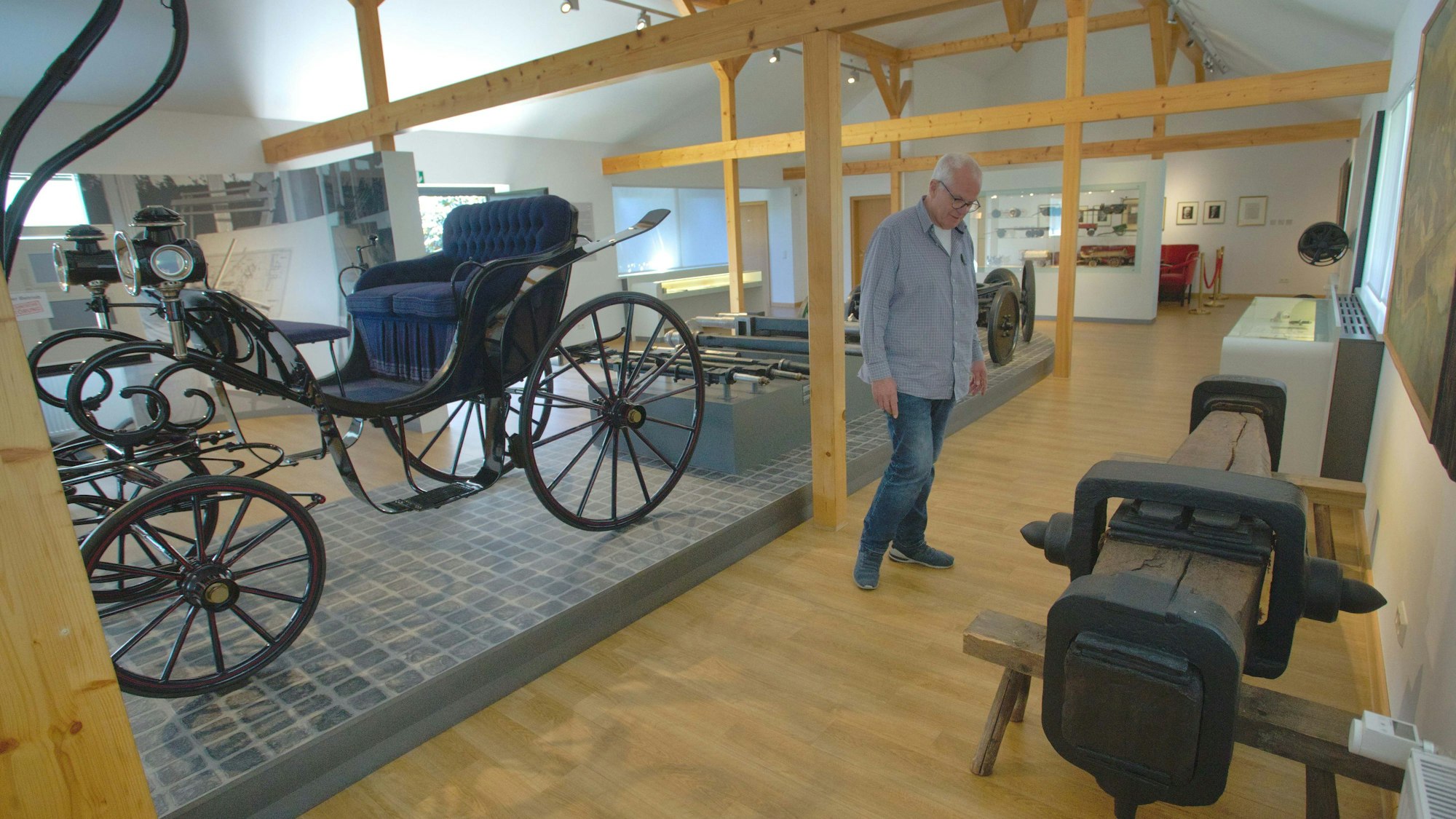 Ein Mann betrachtet ein Fahrwerk aus Holz, dahinter eine Kutsche.