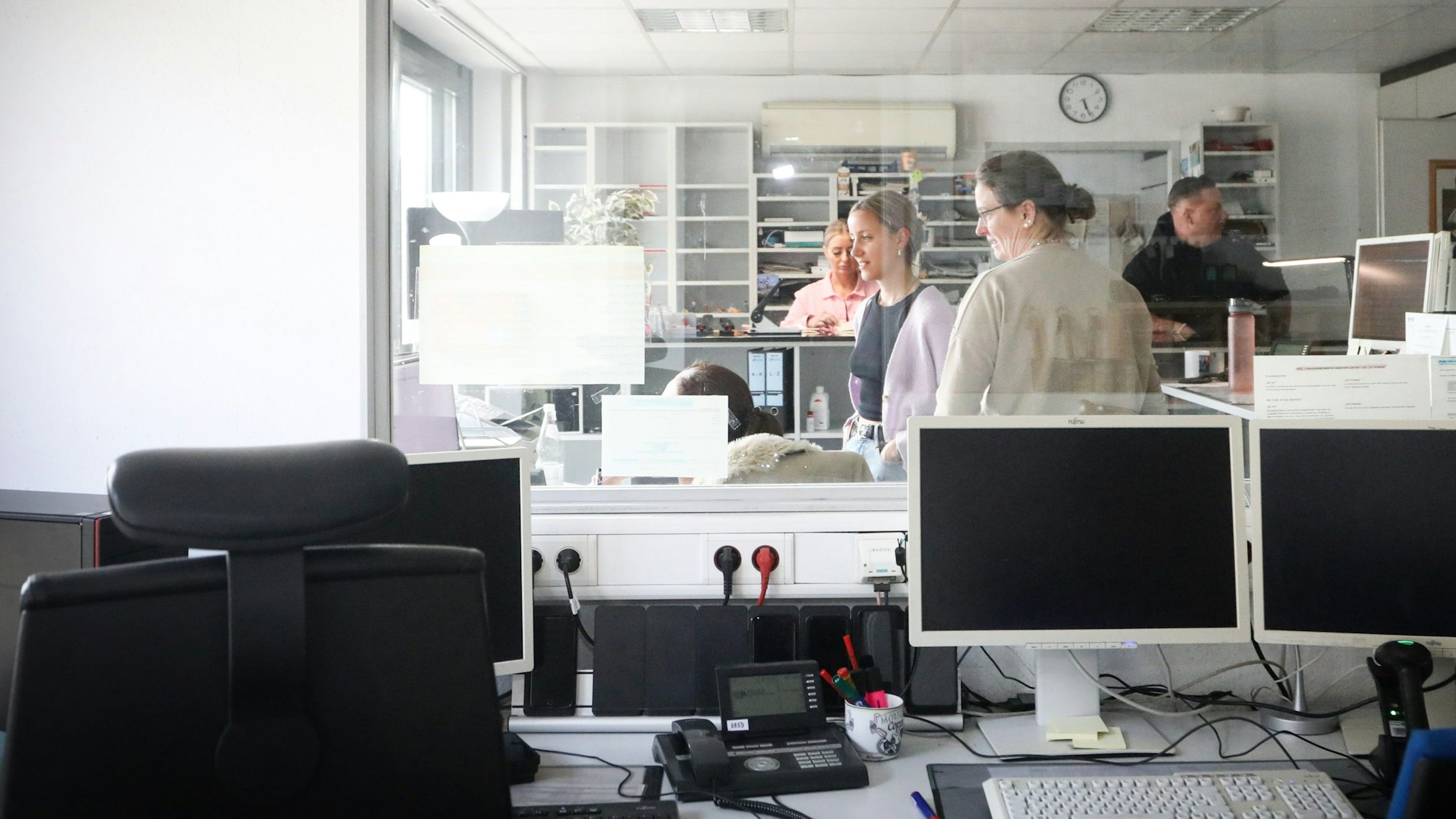 29.03.2025, Köln: Im Vordergrund der Büroräume stehen Computerbildschirme. Hinter einer Scheibe stehen und sitzen Mitarbeitende des Wachsraums der Kriminalwache.