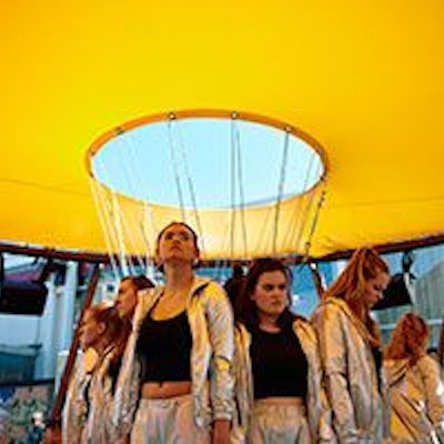 Mädchen in silbernen Trainingsanzügen und schwarzen Tops stehen unter einem gelben Baldachin, der an einer Stahlkonstruktion angebracht ist.