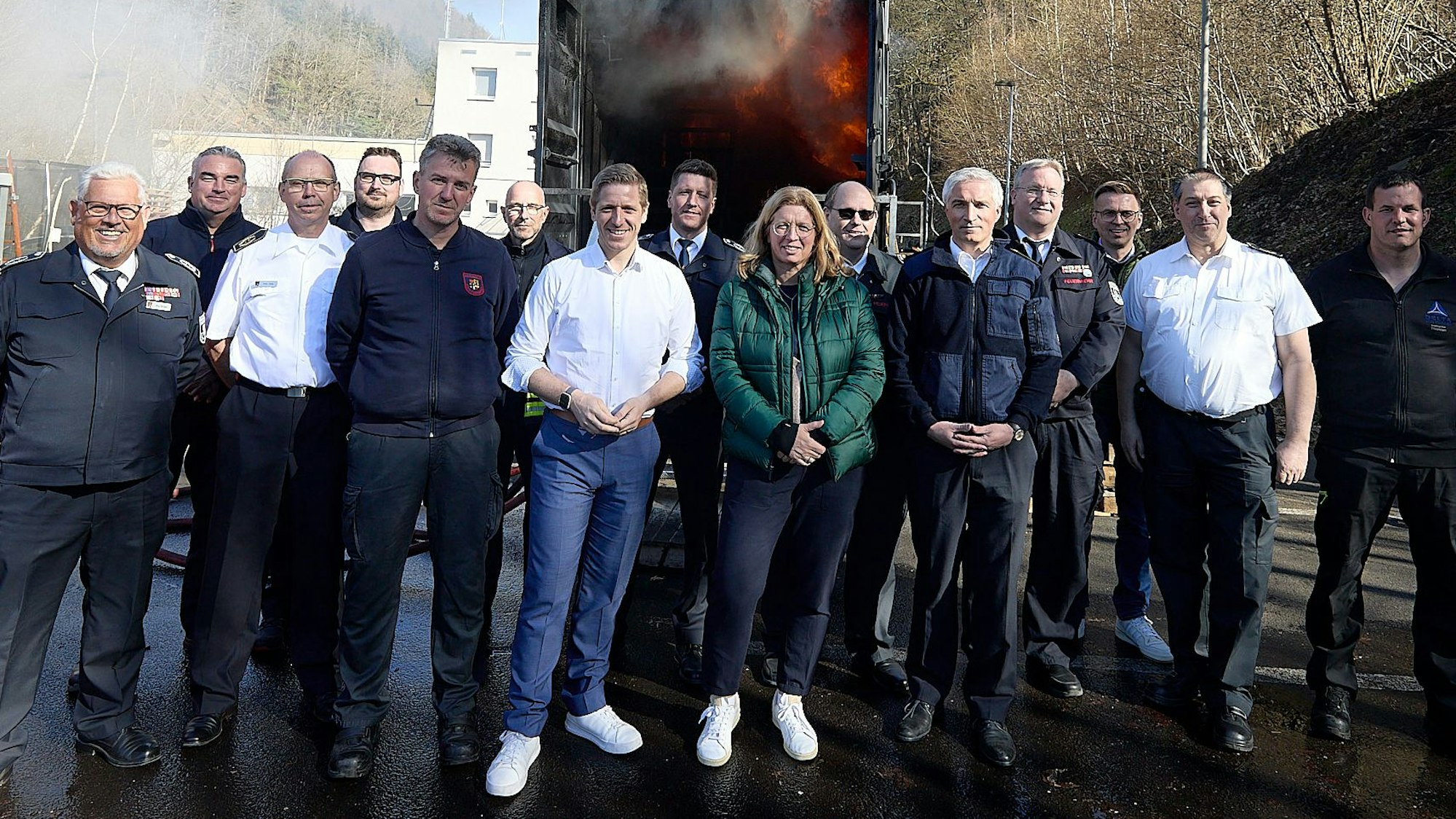 Zahlreiche Männer und eine Frau, einige von ihnen in Feuerwehr-Uniformen, stehen vor dem Container der Brandsimulationsanlage, in dem Feuer zu sehen ist.