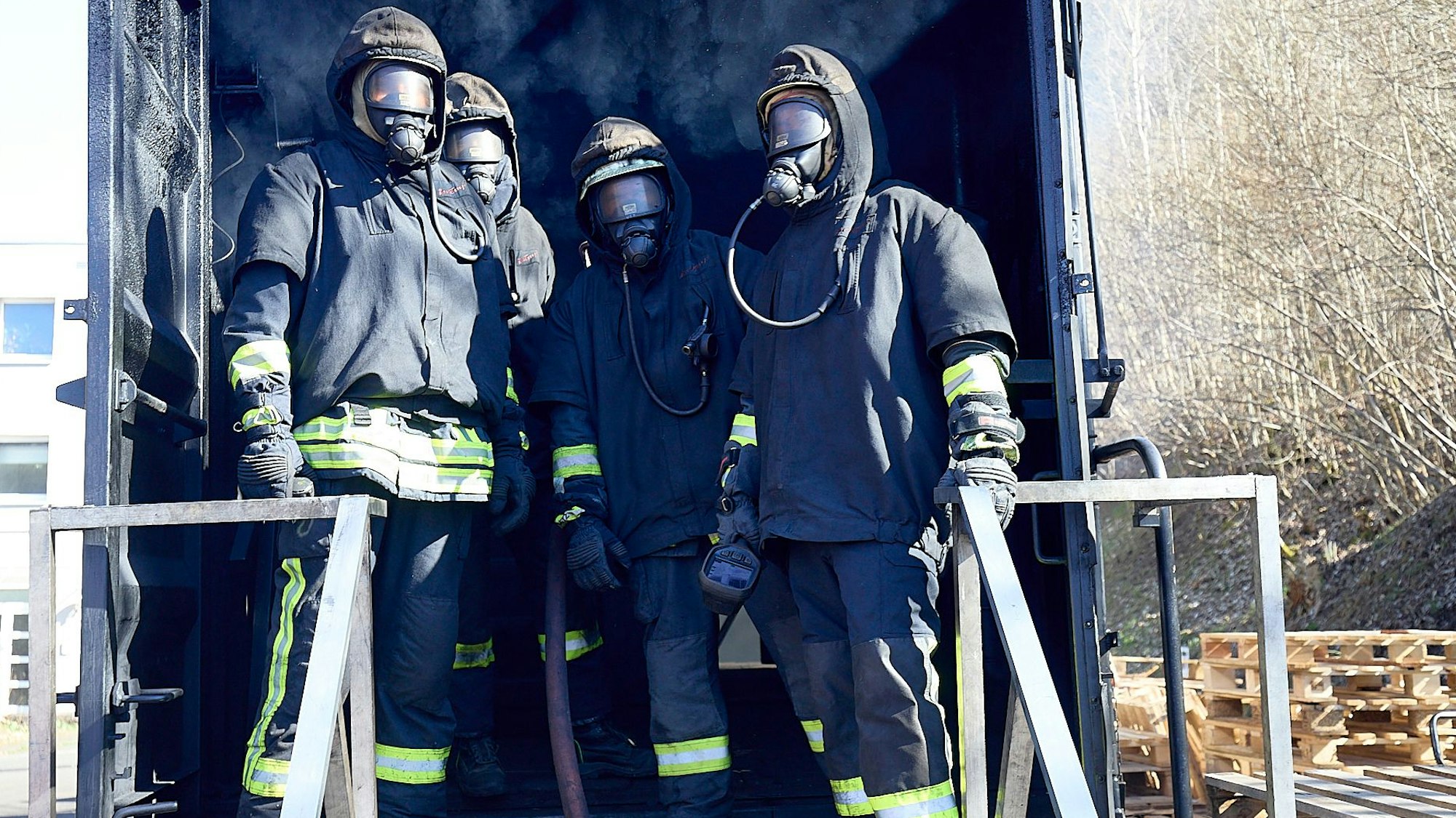 Vier Feuerwehrleute in kompletter Schutzkleidung stehen vor einem Container, aus dem Rauch dringt. Dort absolvieren sie ein Training.