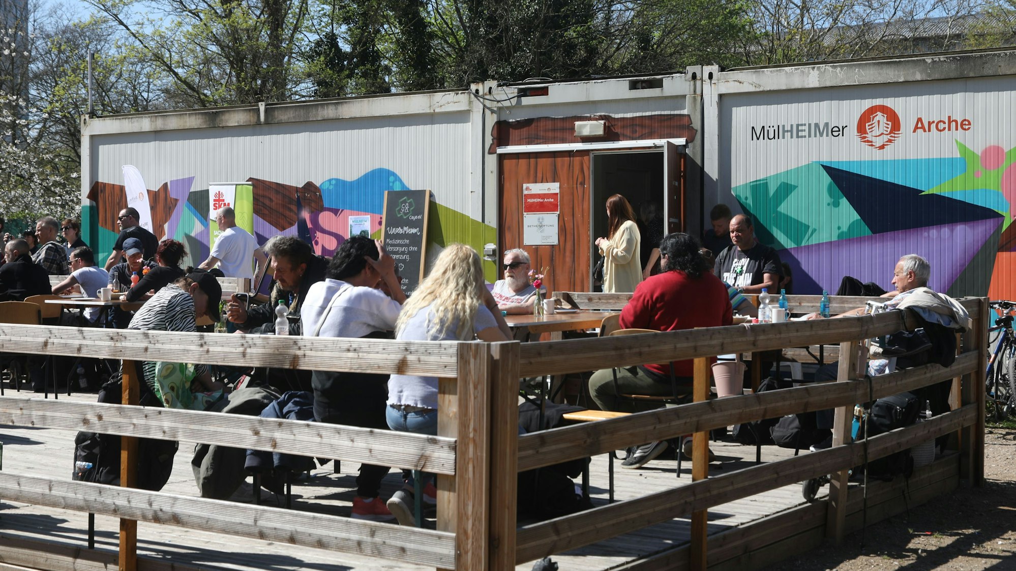 Zu sehen sind Container der Mülheimer Arche am Bergischen Ring. Davor sitzen Menschen an Tischen.