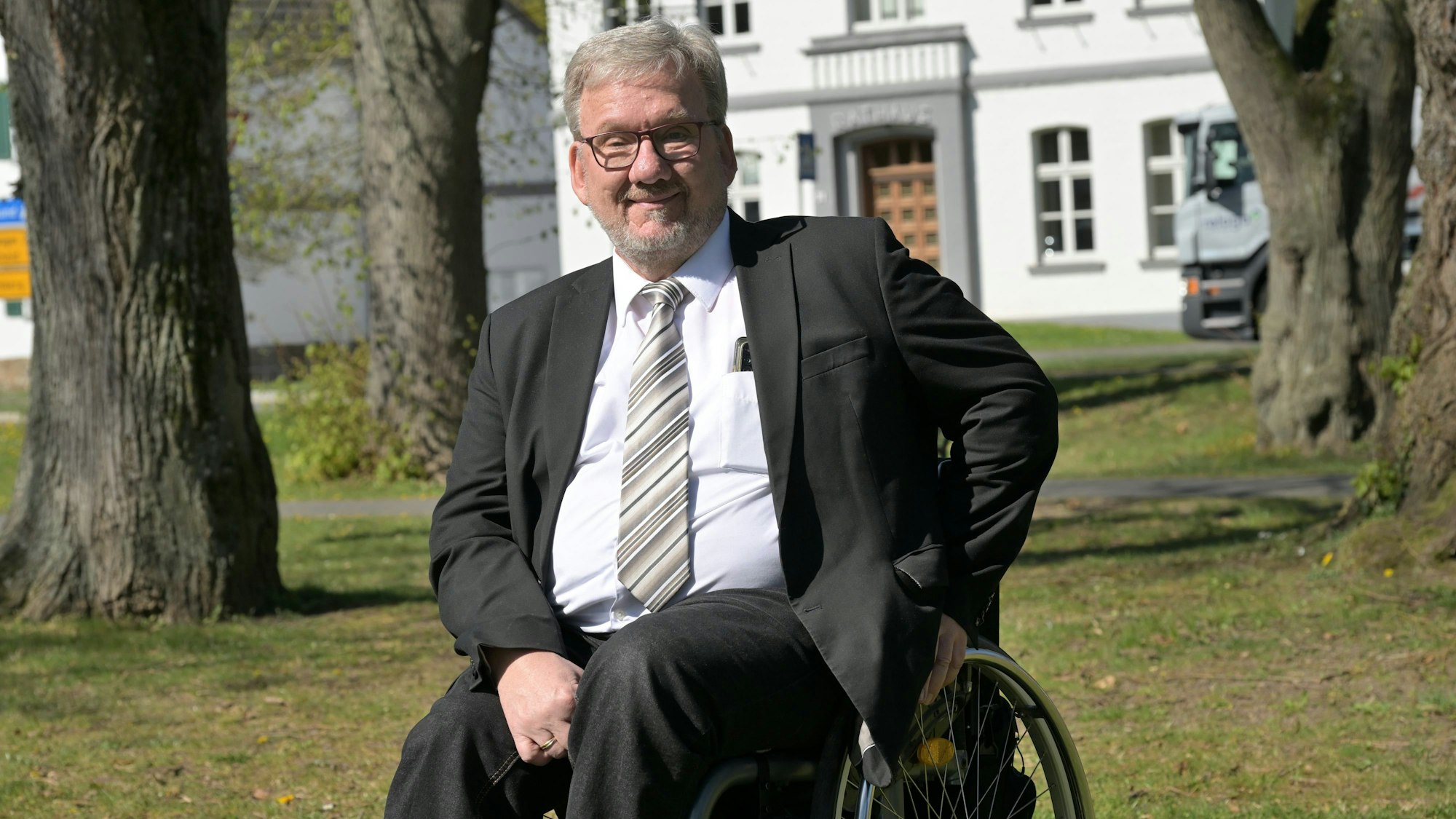 Ein Mann mit Anzug und Krawatte sitzt im Rollstuhl. Hinter ihm ein Rathaus.