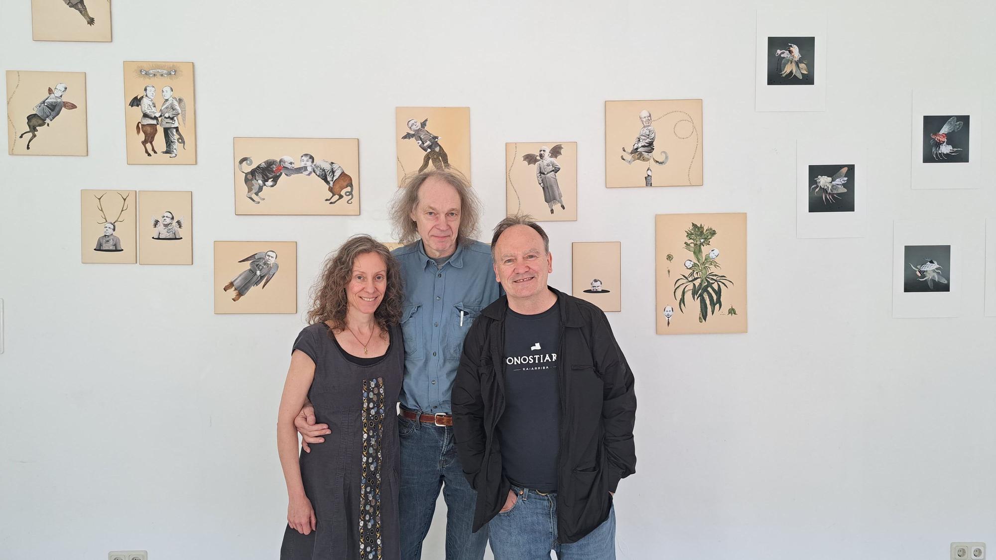 Eine Frau und zwei Männer stehen nebeneinander vor einer Wand, an der Bilder hängen.