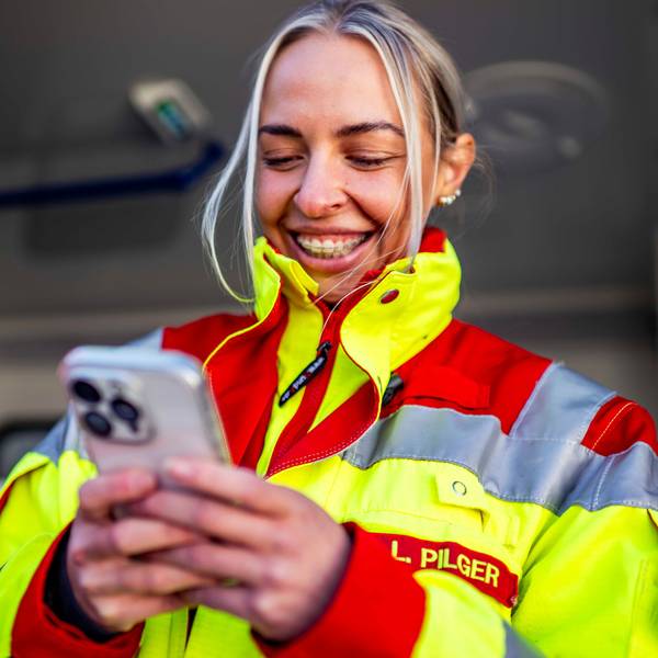Das Bild zeigt Lea Pilger in ihrer Rettungsdienstkleidung. Sie hält ein Smartphone in der Hand hat.