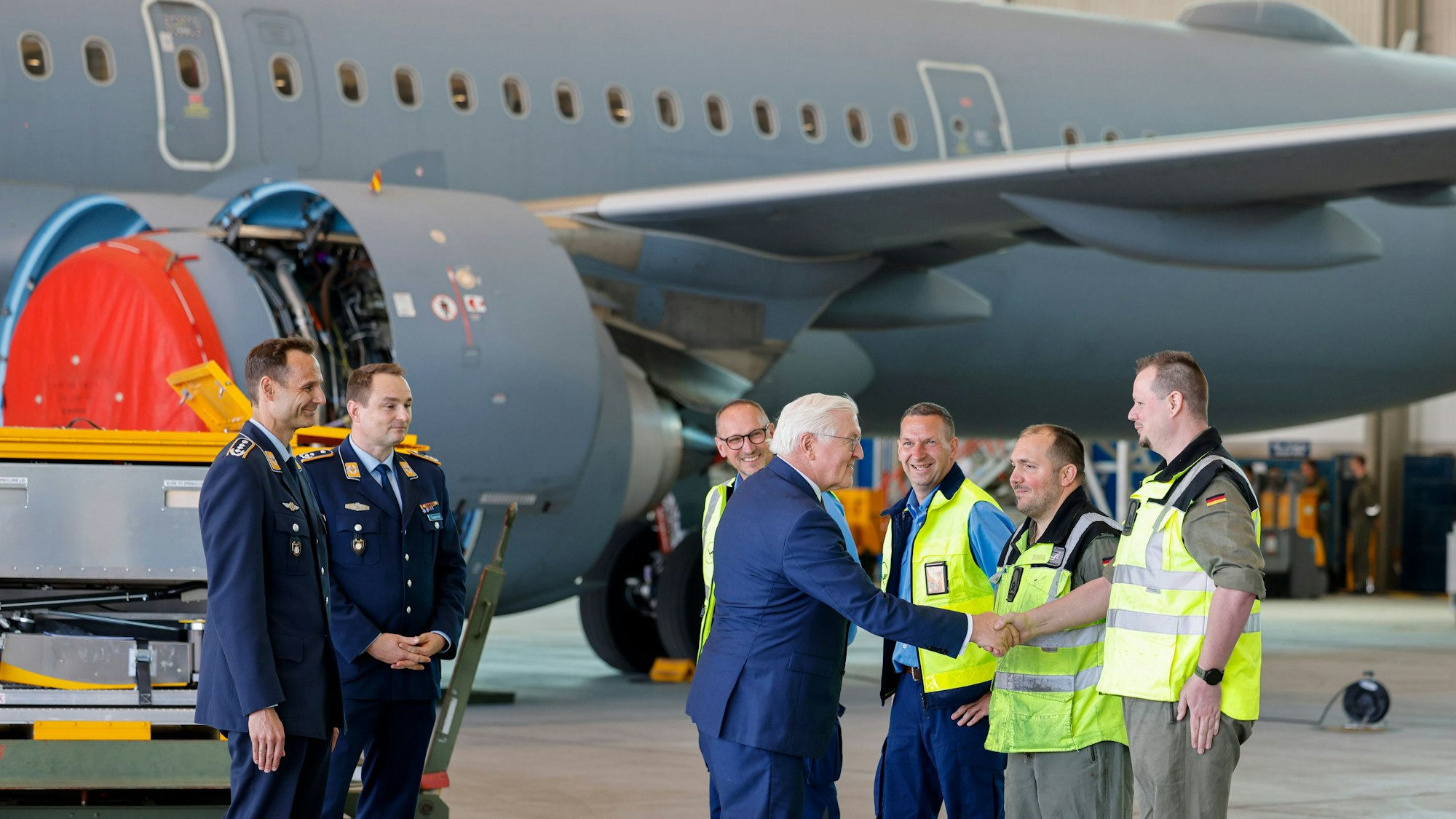 Zu sehen ist Frank-Walter Steinmeier in der Seitenansicht vor einem Flugzeug stehend. Er begrüßt vier Personen in gelben Westen. Hinter ihm stehen zwei Offiziere.