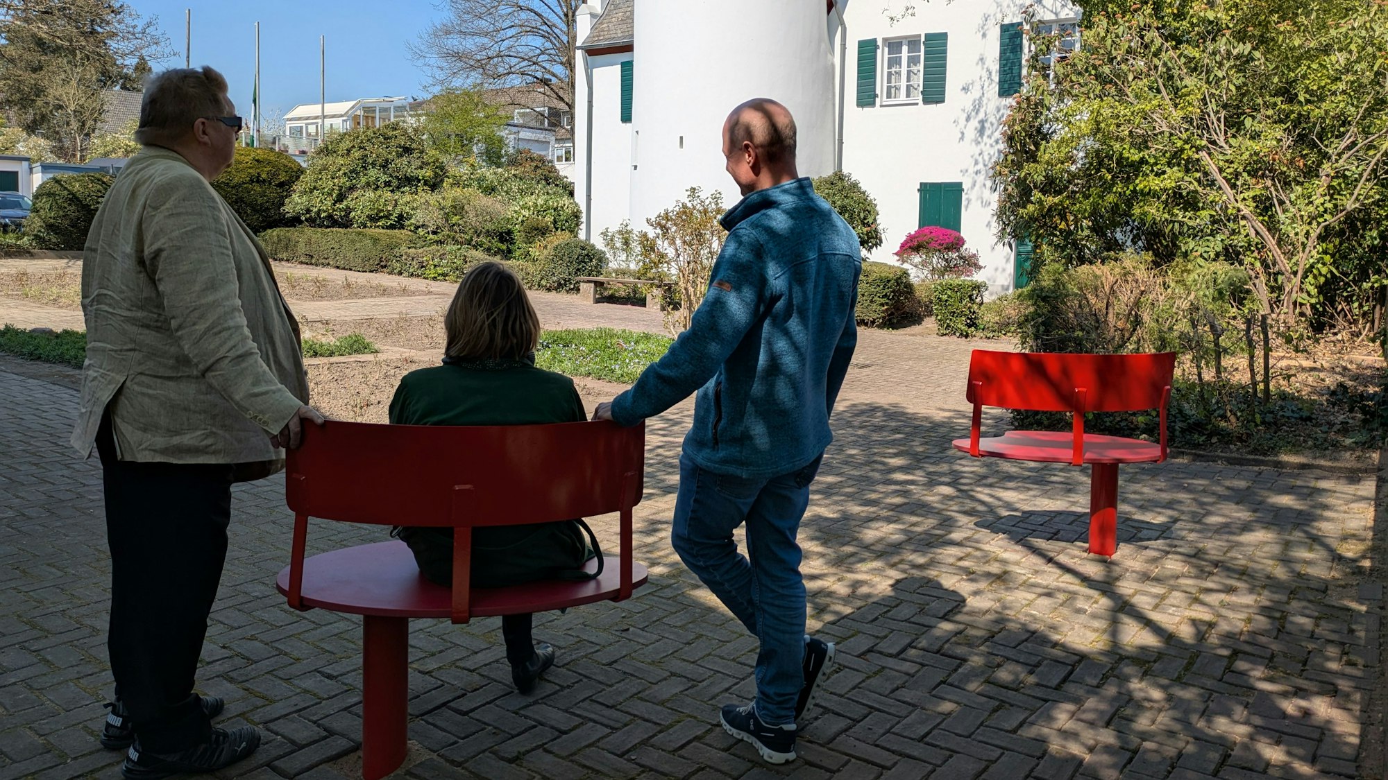 Drei Personen blicken von einer roten Bank aus auf ein neues Blumenbeet.