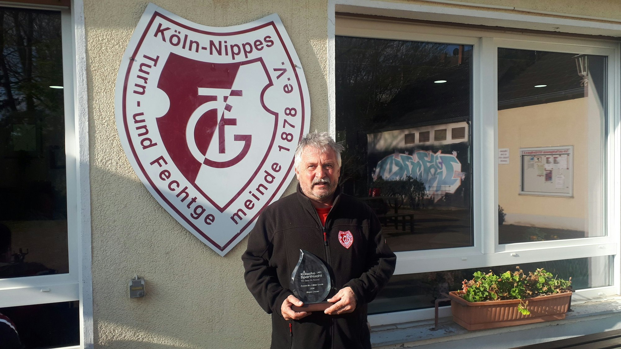 Ein Mann mit grauem Haar und Schnauzer hält eine Trophäe in der Hand. Dahinter ist an einer Hauswand ein Vereinsschild zu sehen, auf dem Köln-Nippes, Turn- und Fechtgemeinde 1878 e. V. steht.