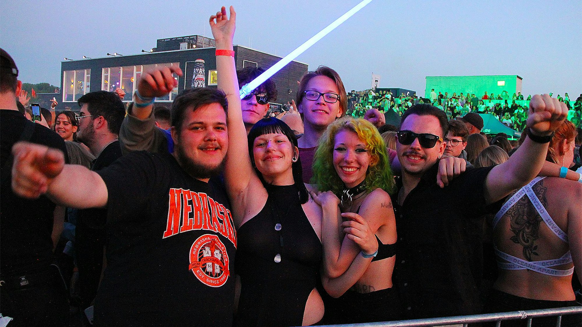 Junge Menschen in dunkler Kleidung stehen in einer Menge auf einem Konzertgelände. Sechs Personen posieren für ein Gruppenfoto.