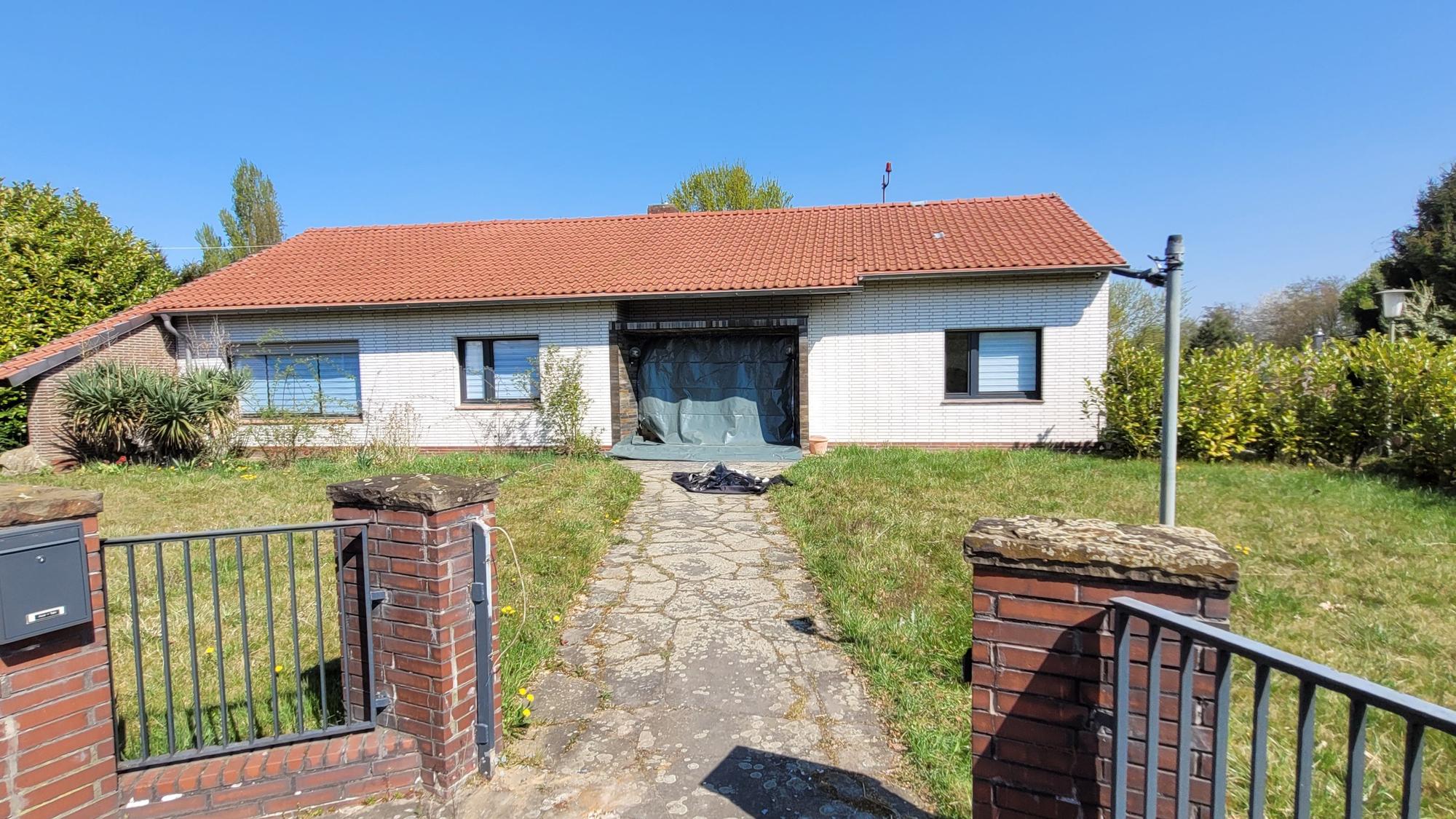 Ein in die Jahre gekommenes Haus mit heller Fassade, die Rolläden an den drei Fenstern sind heruntergelassen, dunkle Folie ersetzt die Eingangstür.